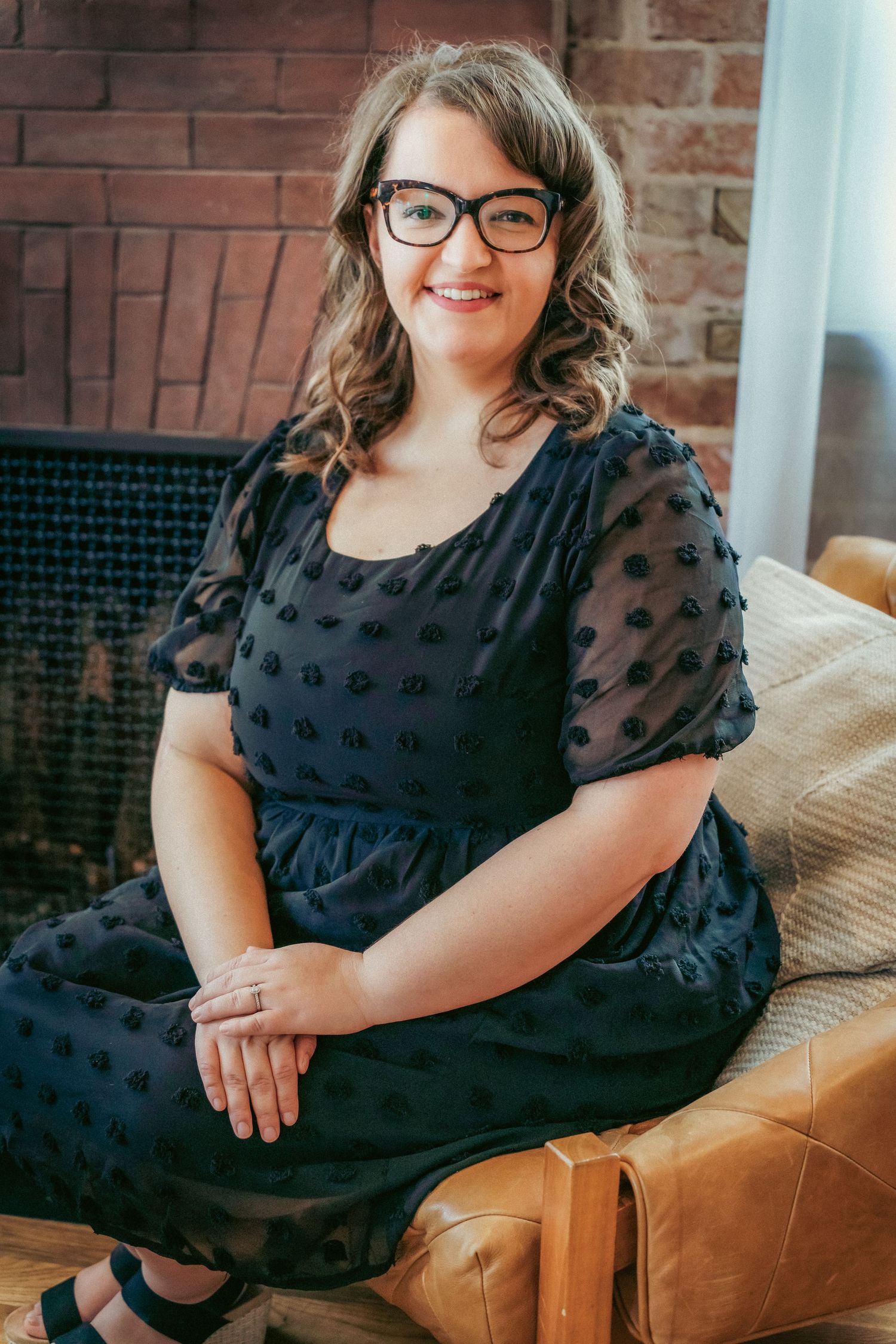 A person in a black dress sits on a brown leather couch against a brick wall.