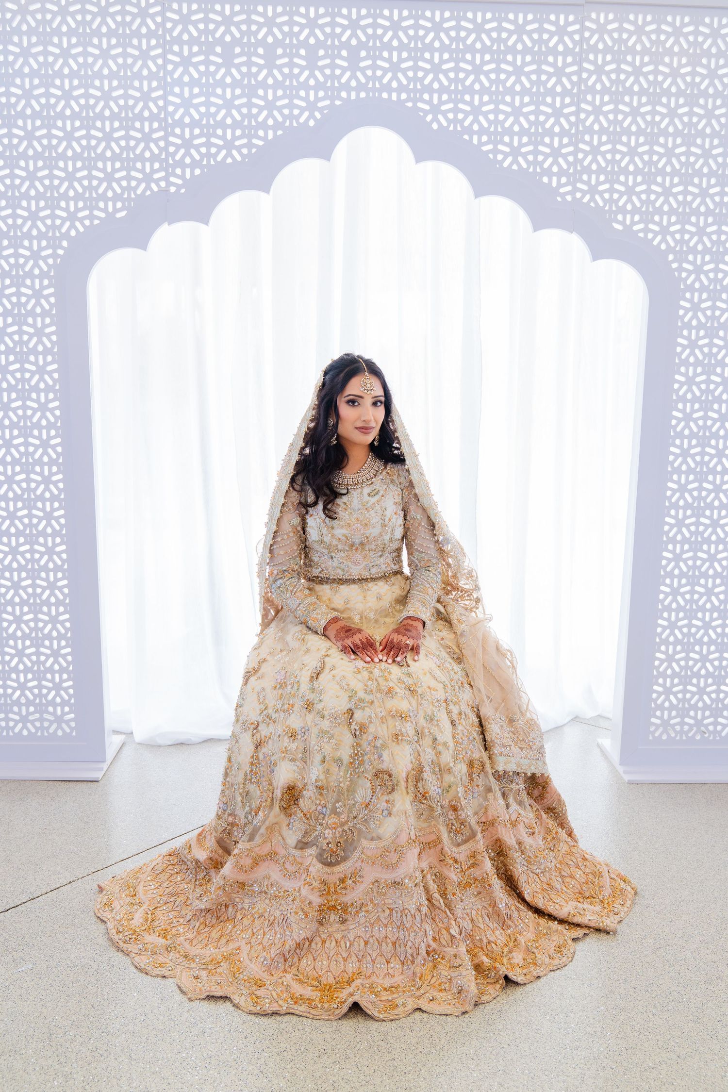 Traditional bride in cream lehenga poses gracefully against white geometric backdrop.