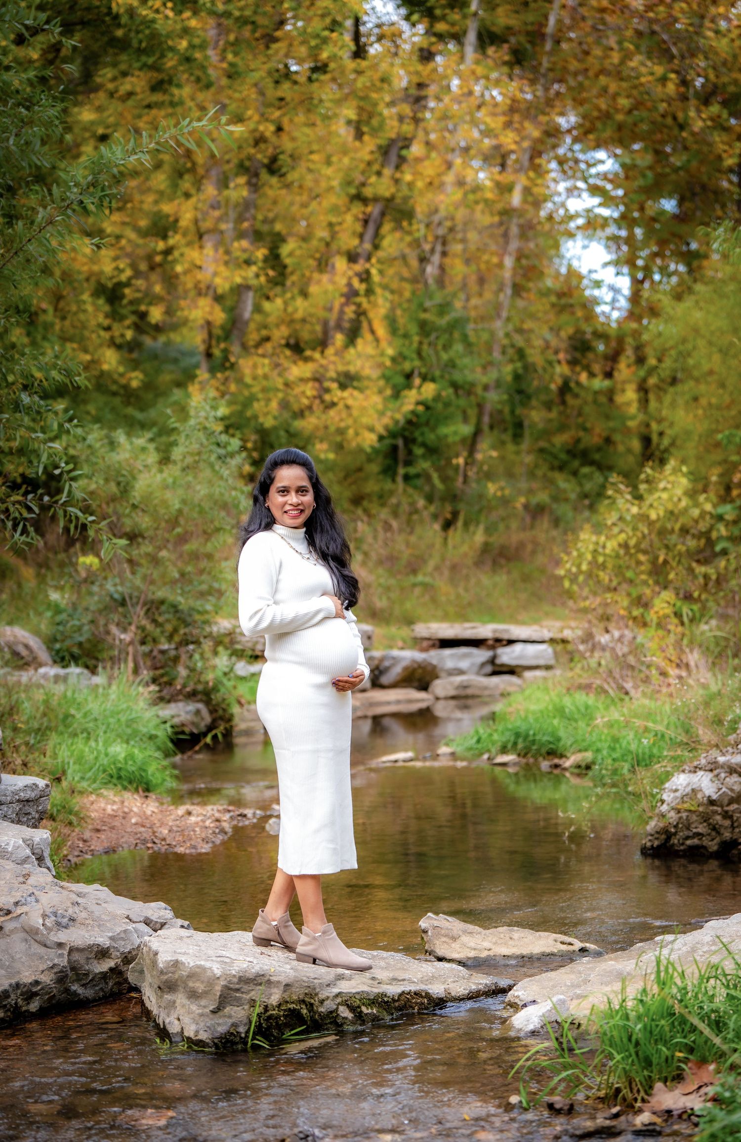 A woman in a white dress stands on rocks near a peaceful stream surrounded by autumn foliage.