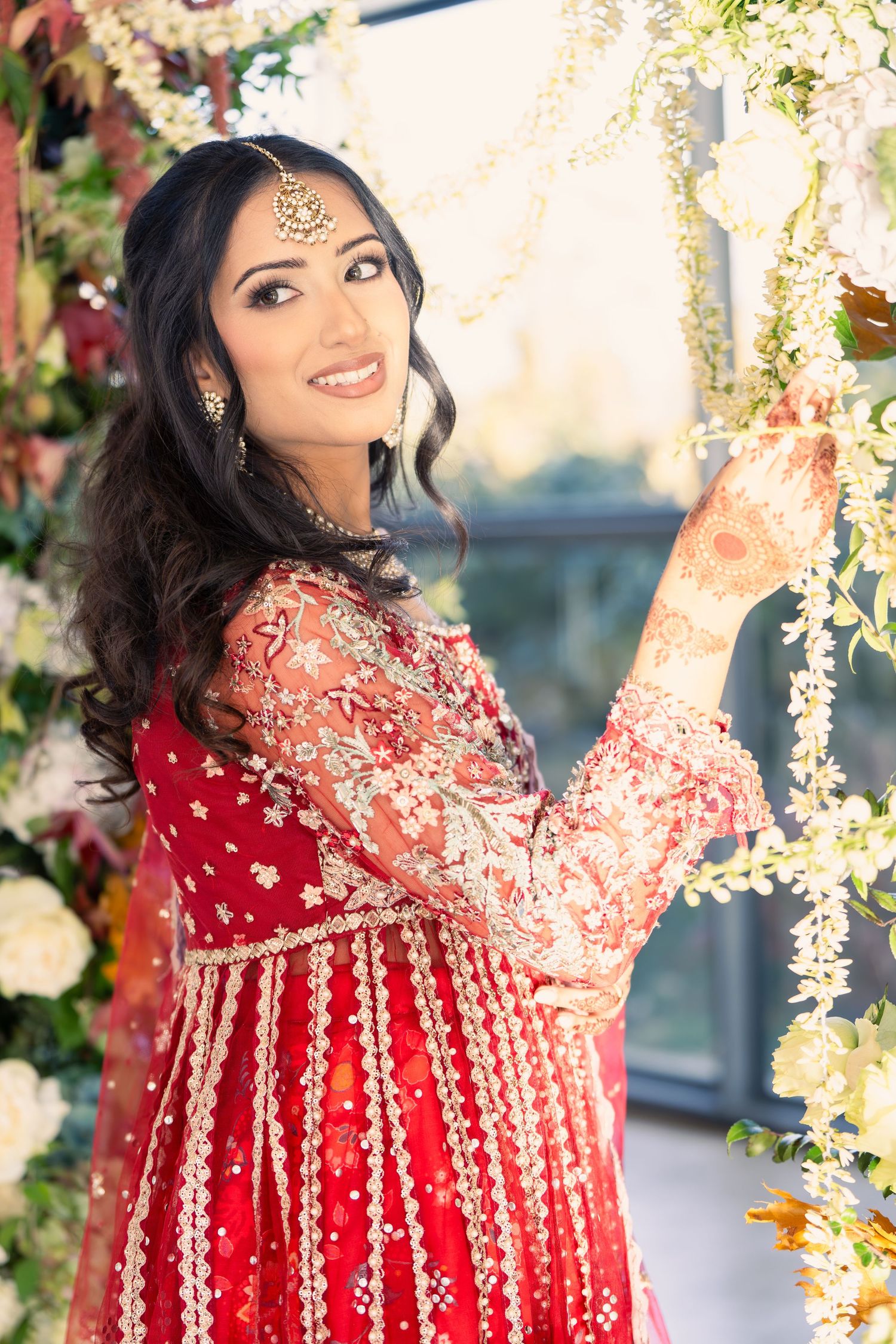 Bride in red traditional Indian wedding attire posing against floral backdrop.