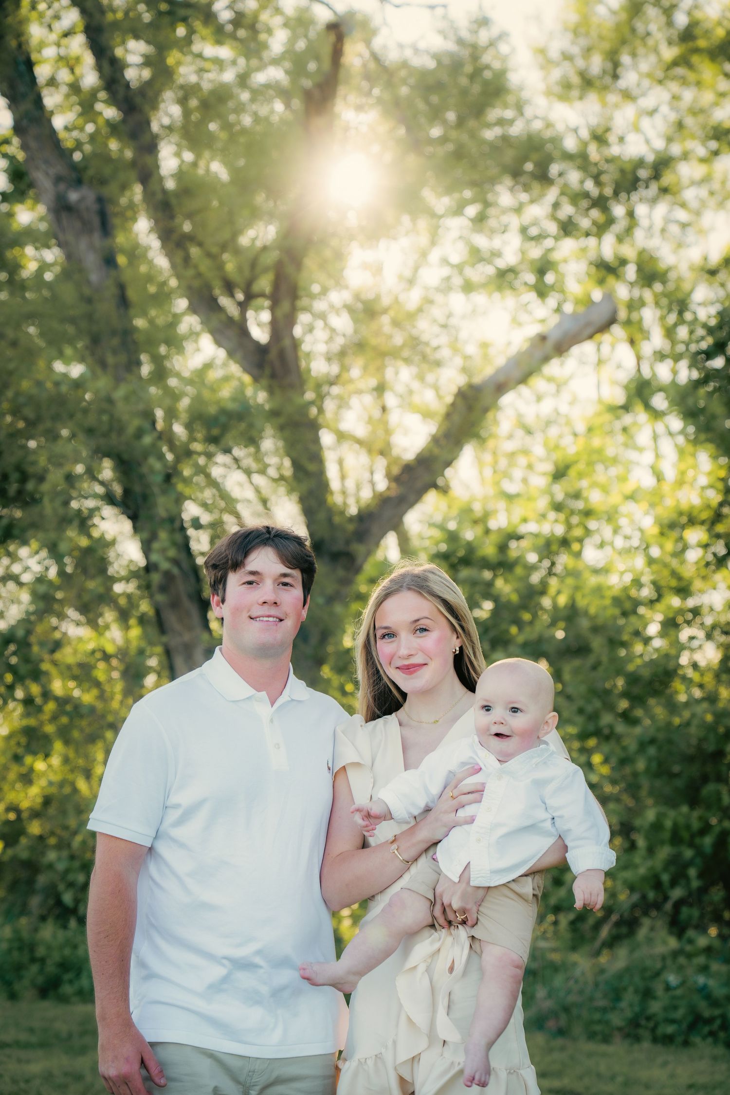 Family portrait in white attire against sunlit trees in a park setting.