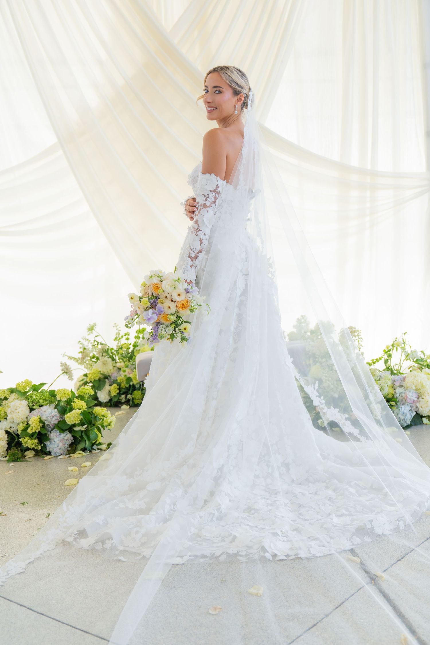 Full-length wedding dress photo showing intricate lace train and veil against white backdrop.
