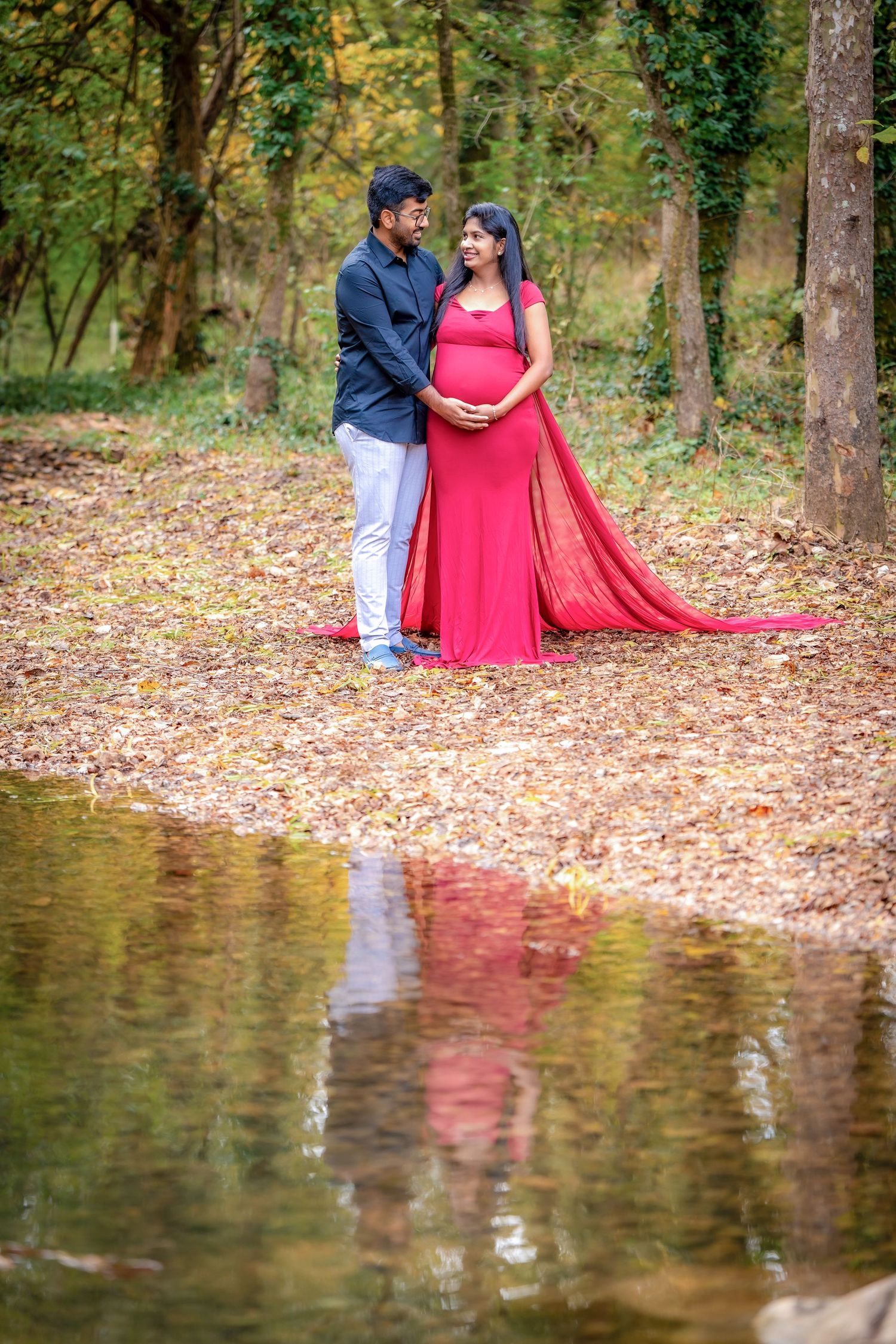 A couple poses romantically by a reflective pond surrounded by fallen autumn leaves in a wooded setting.