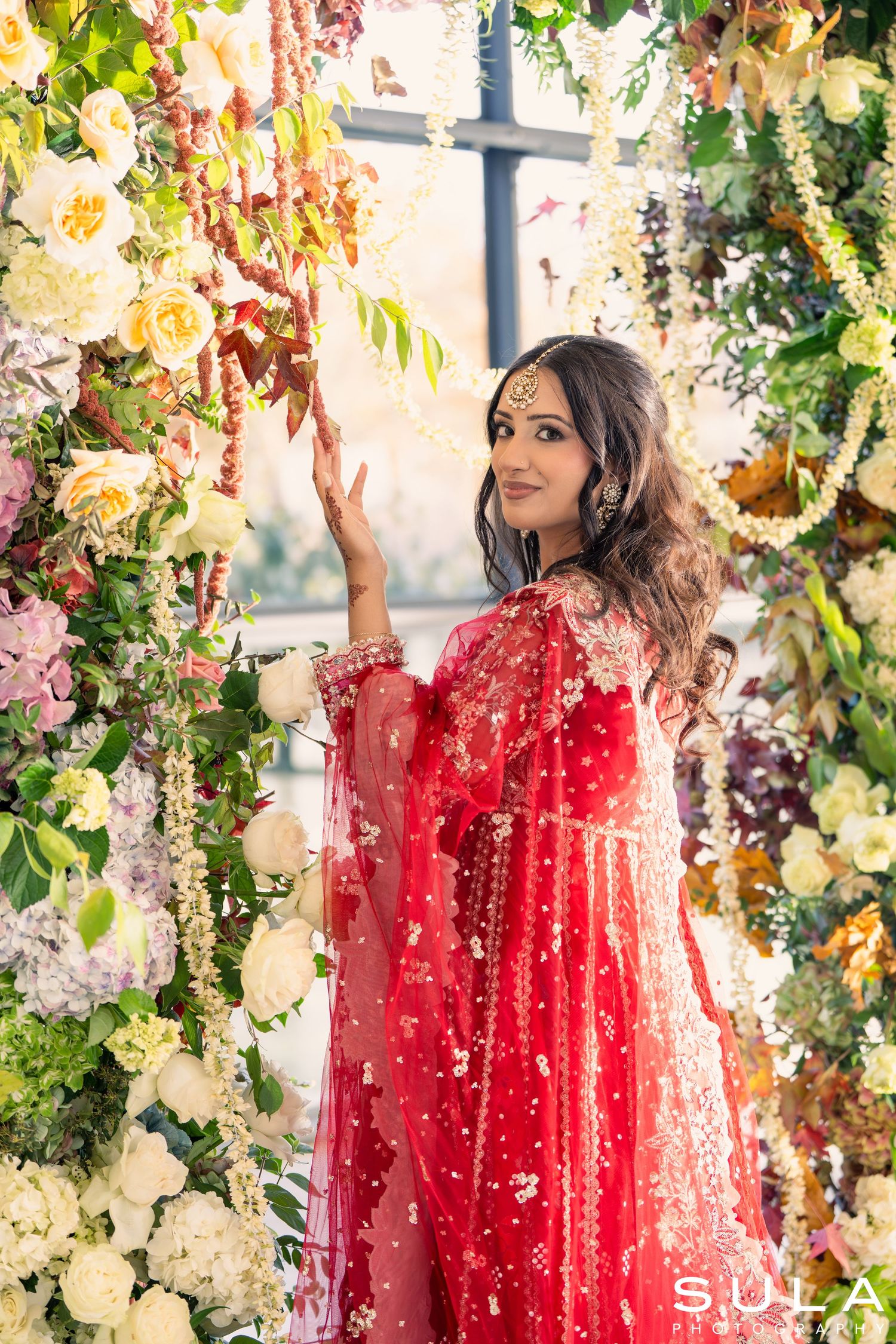 Red embellished dress photographed in a lush garden setting with colorful floral arrangements.