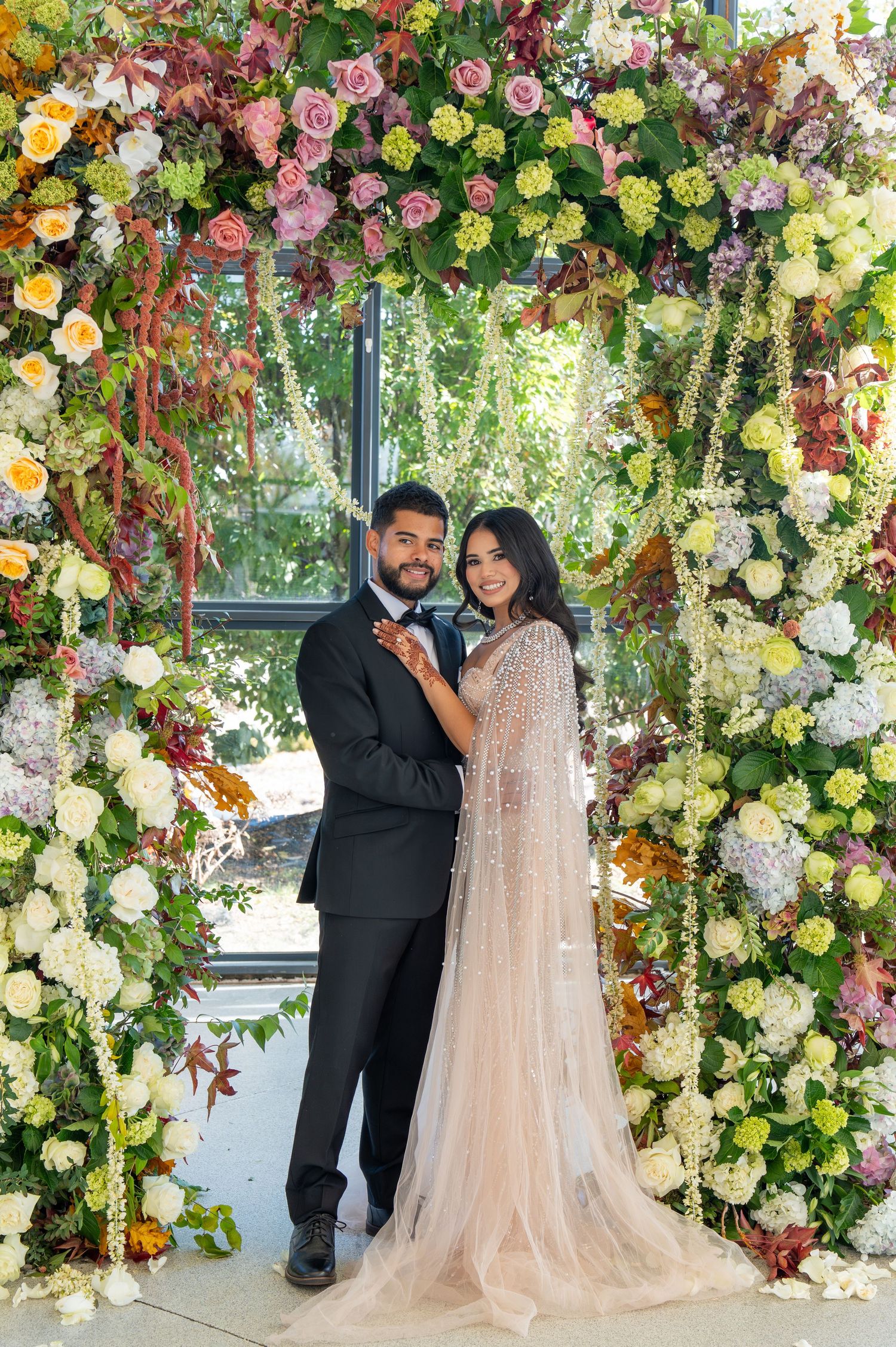 A romantic wedding portrait under a lush floral arch with pink and yellow roses and cascading greenery.