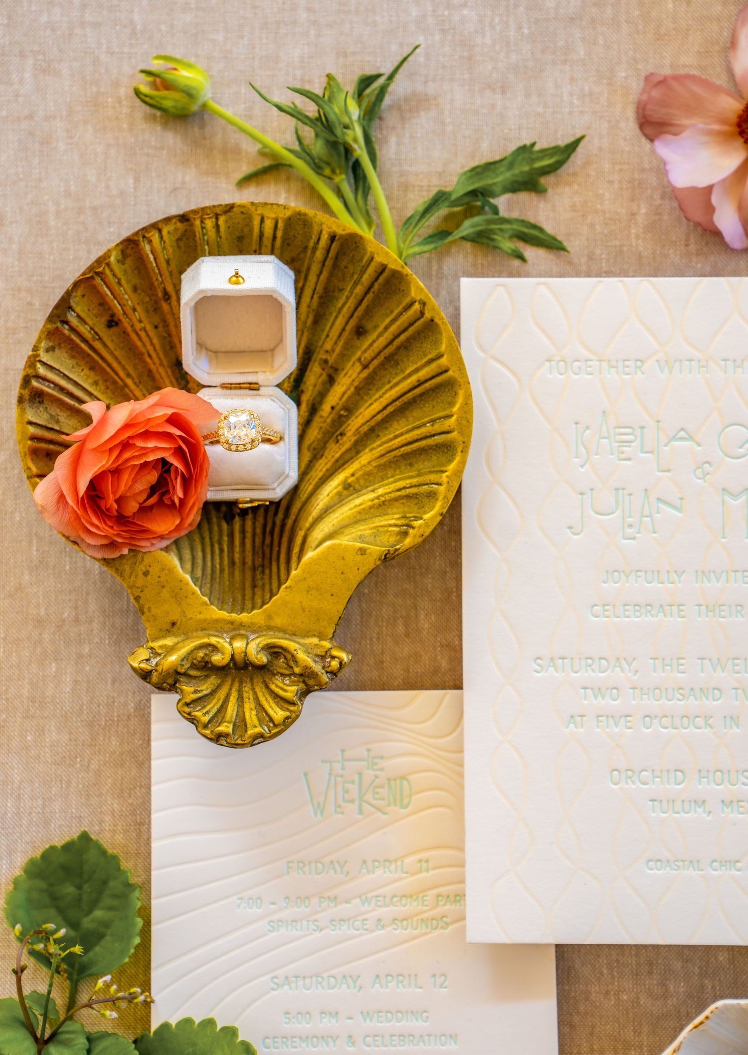 Gold shell-shaped ring holder with coral rose and wedding invitation on linen background.