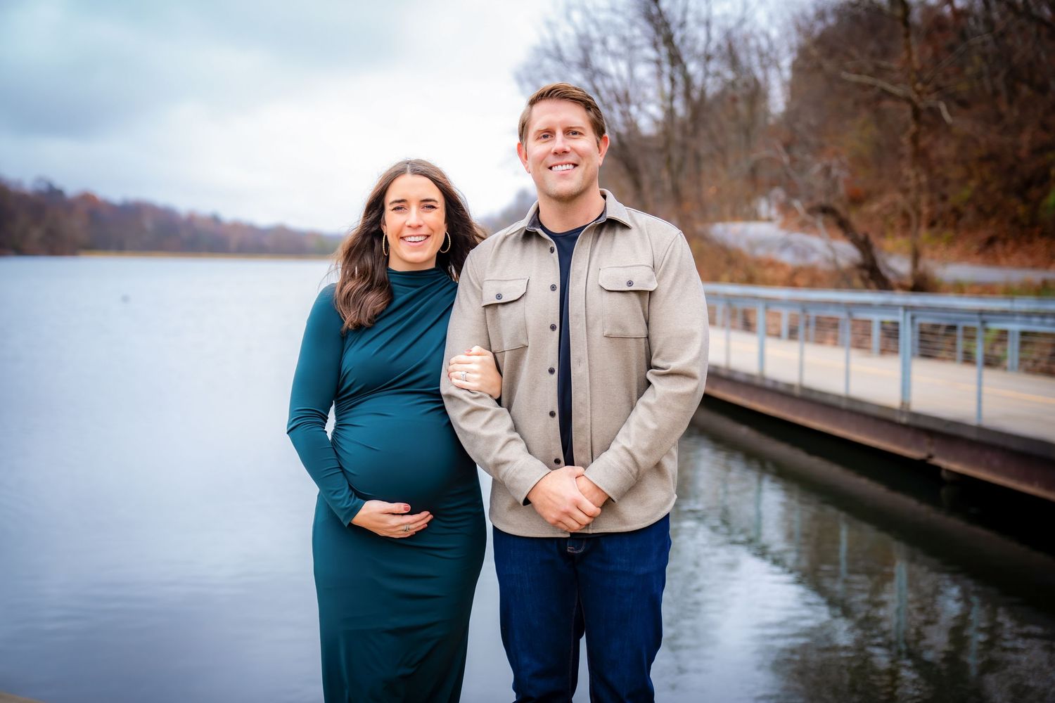 Couple posing by a lake with bare winter trees and cloudy skies in the background.