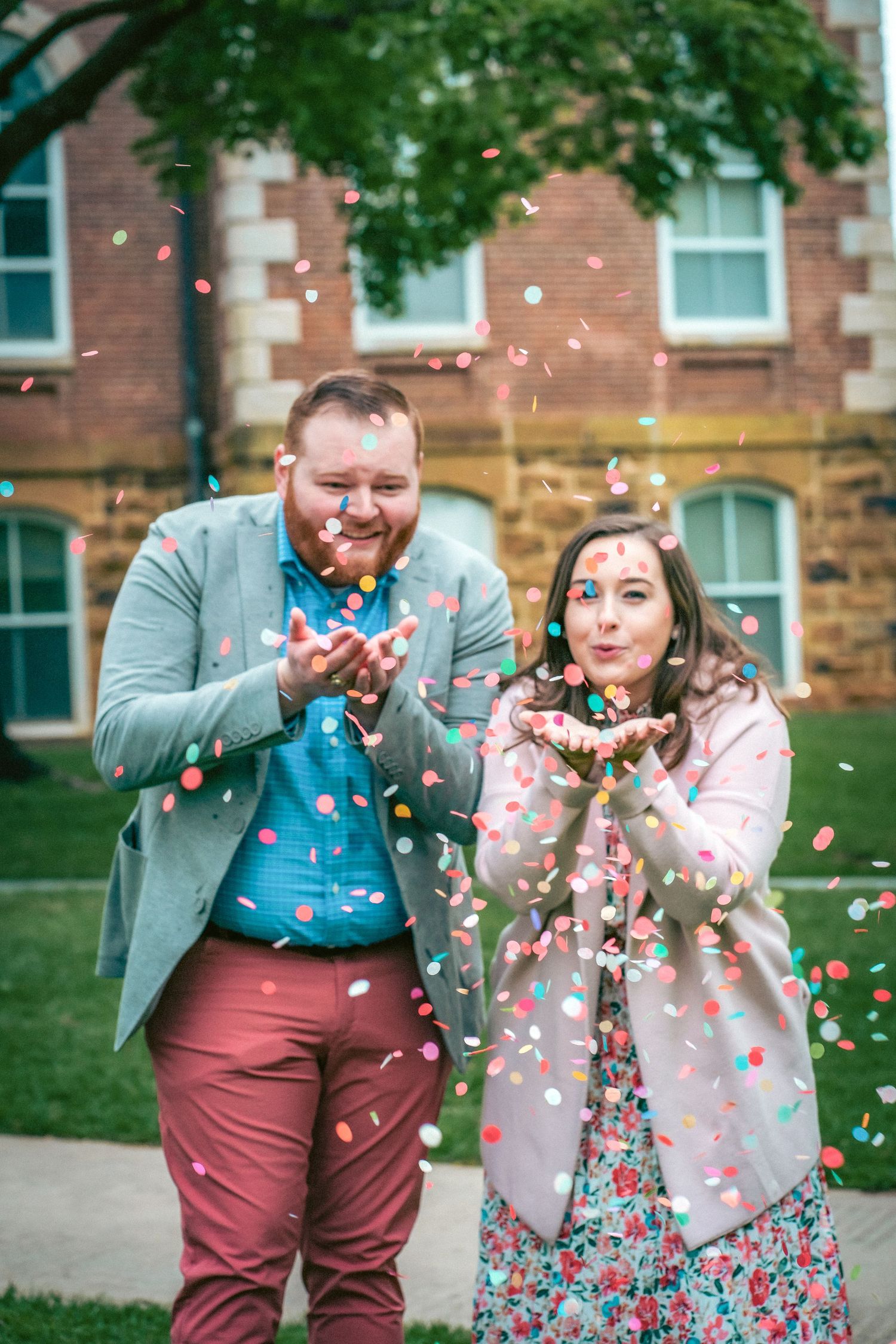 Couple celebrating with colorful confetti toss in front of collegiate architecture.