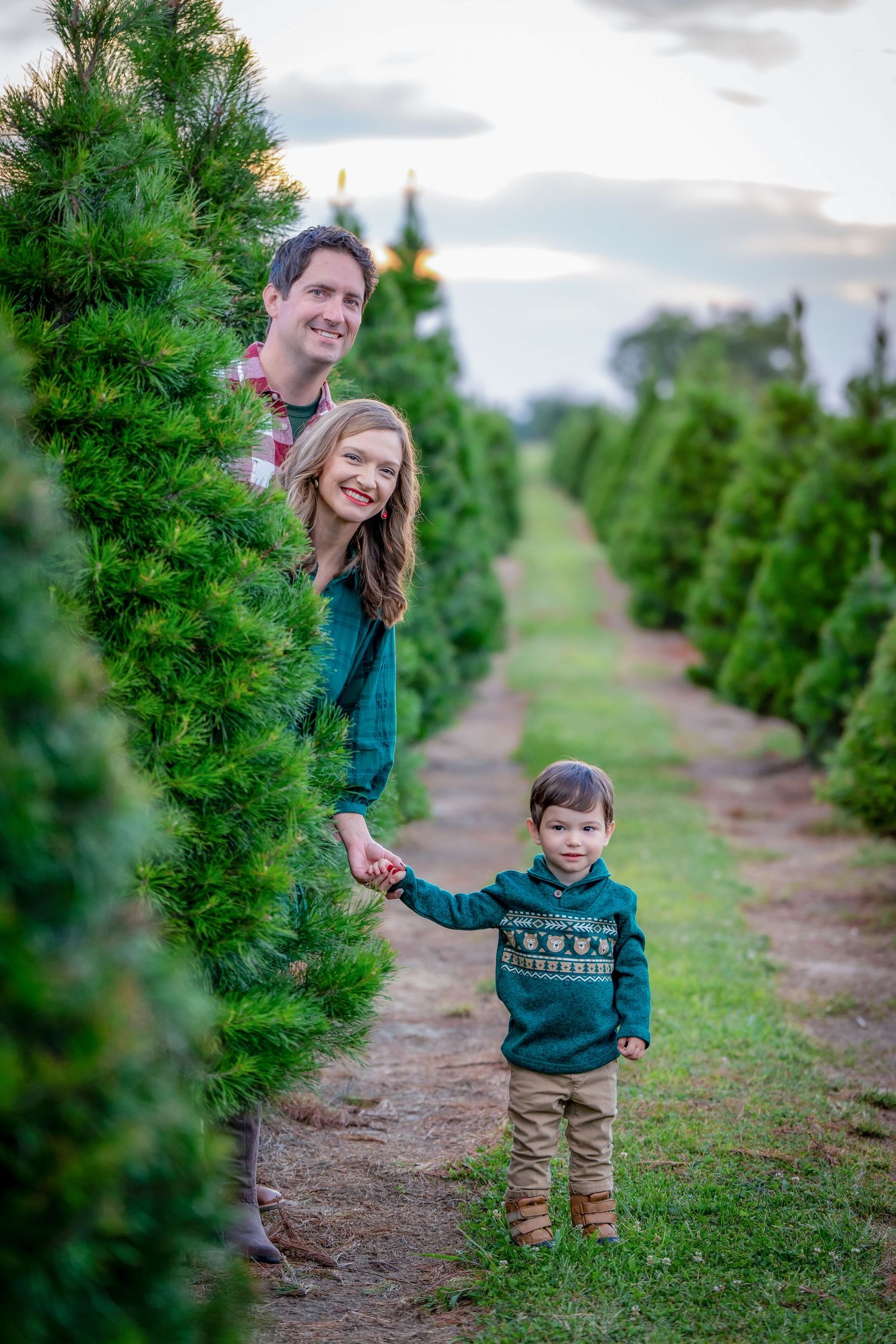 Family walking through rows of evergreen trees at a Christmas tree farm on a cloudy day.
