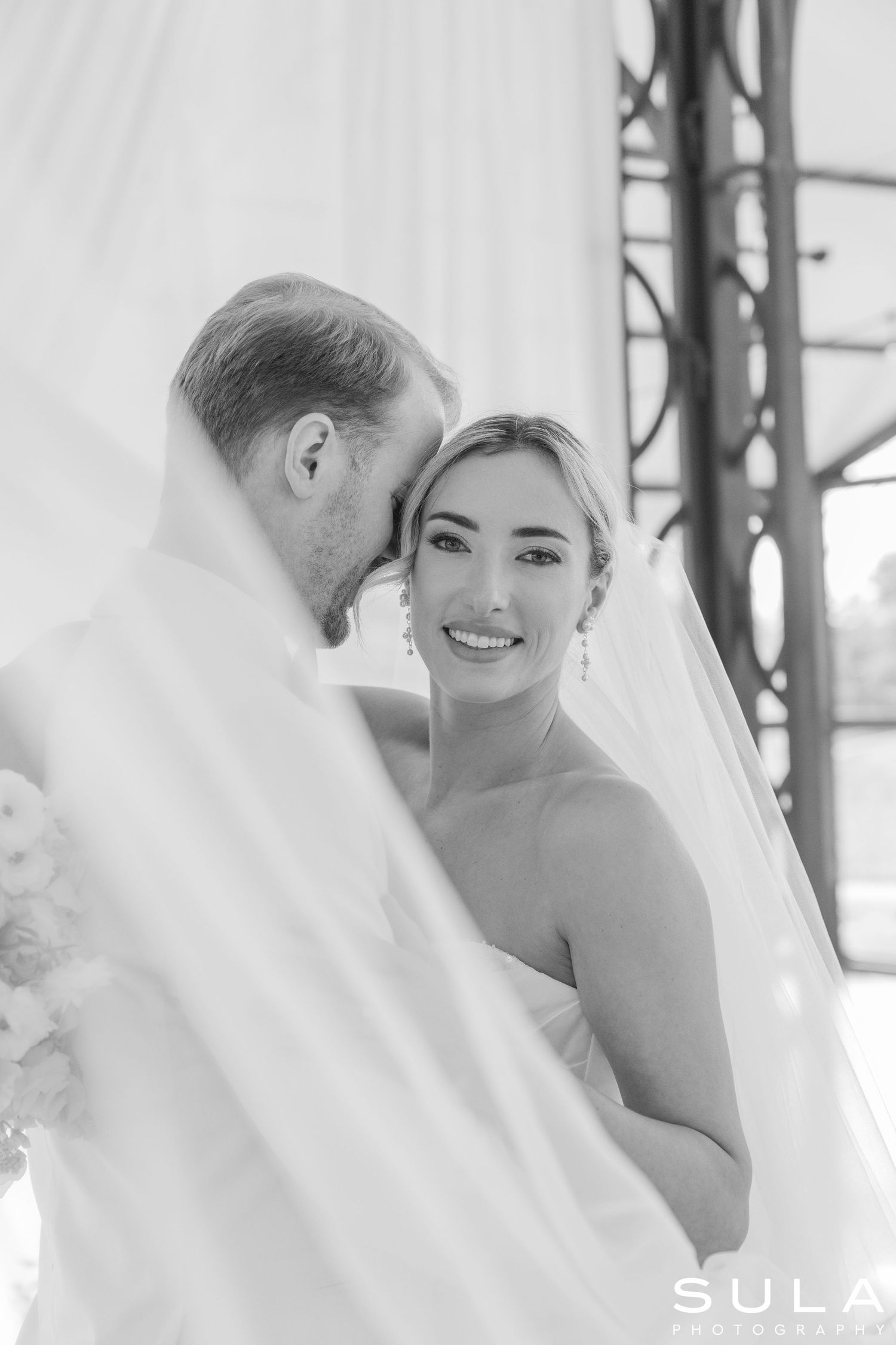Black and white wedding photograph showing a tender moment between newlyweds under a veil.