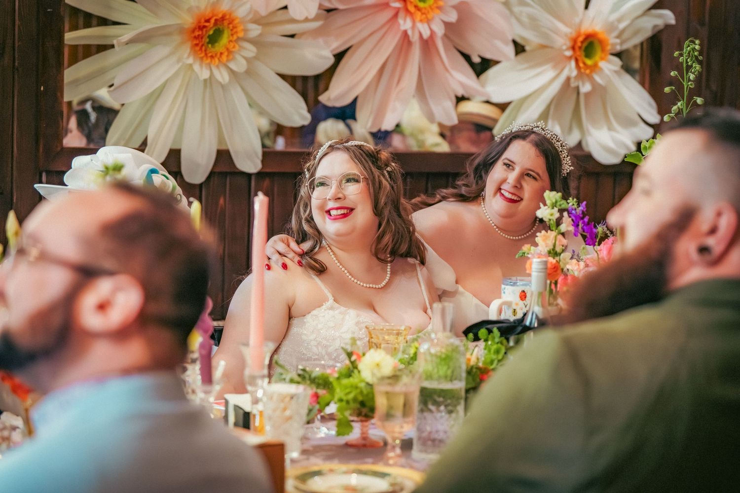 Wedding reception guests laugh joyfully at a table decorated with oversized daisy flowers overhead.