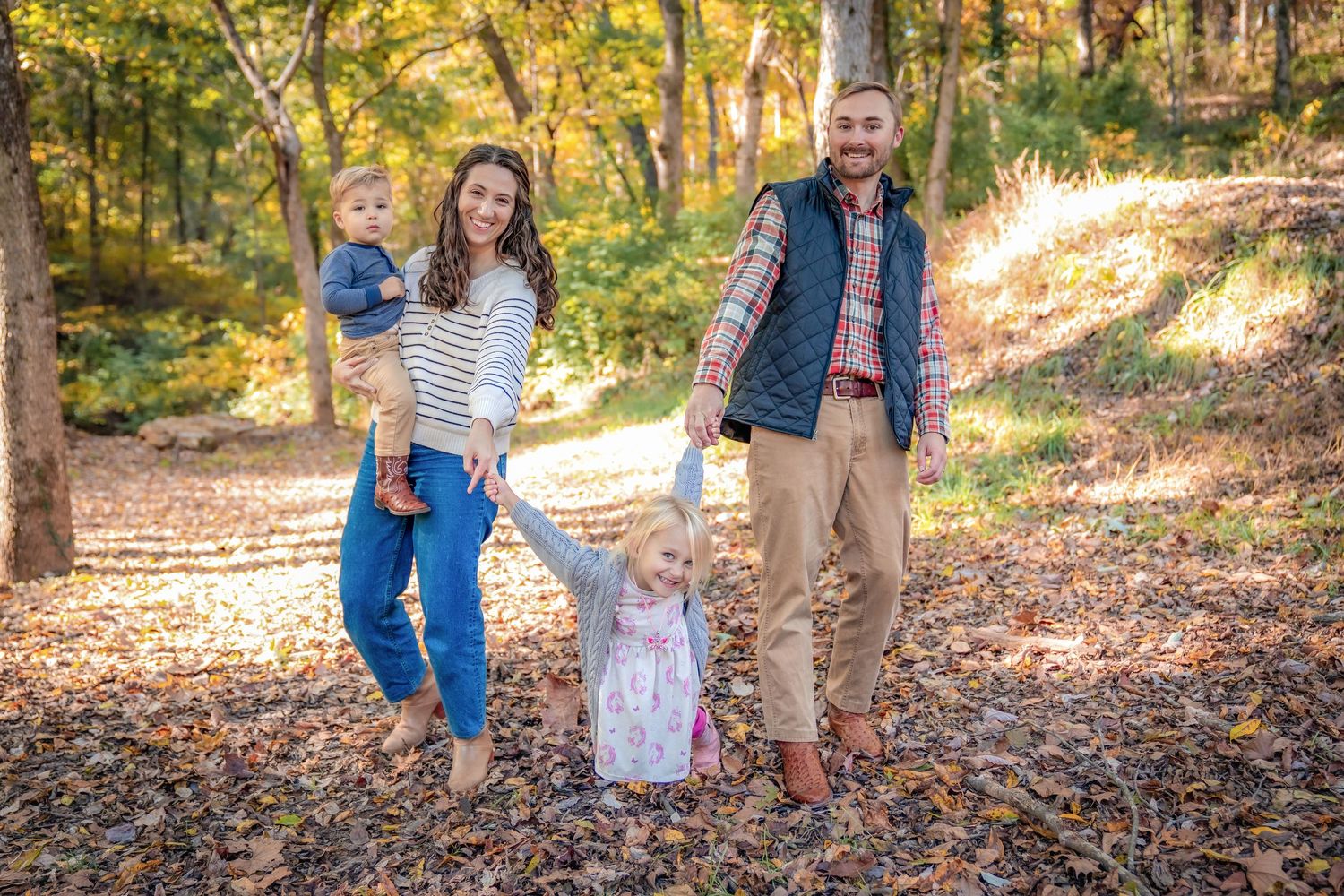 Family walking together on a leaf-strewn path through golden autumn woods.