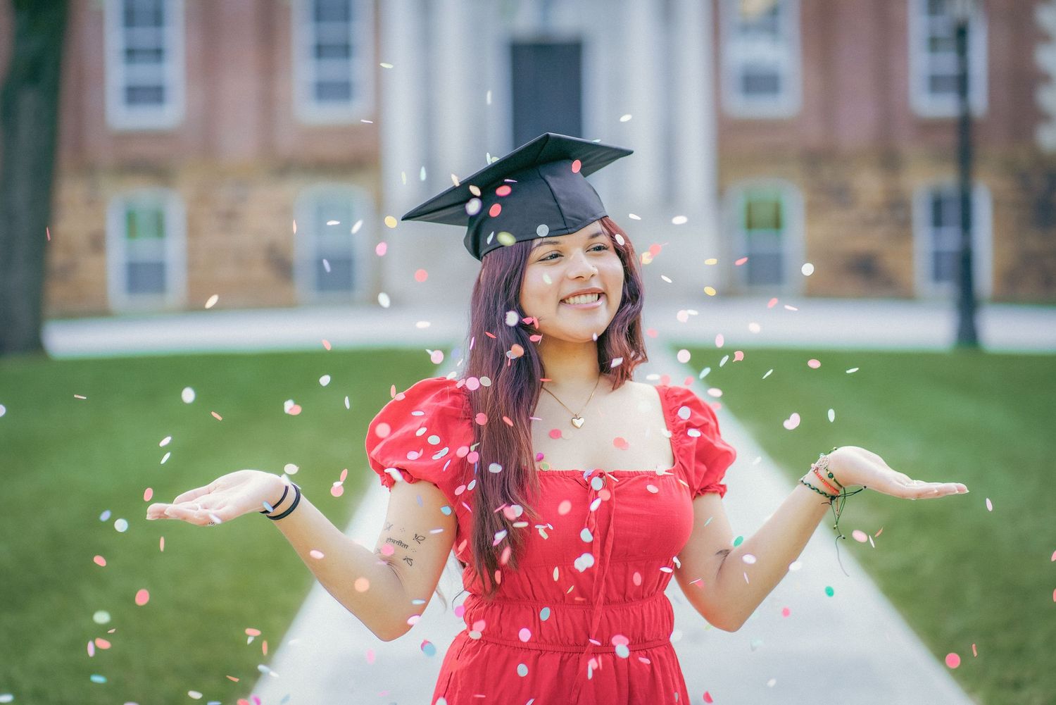 Graduate celebrating with confetti toss while wearing red dress and cap.