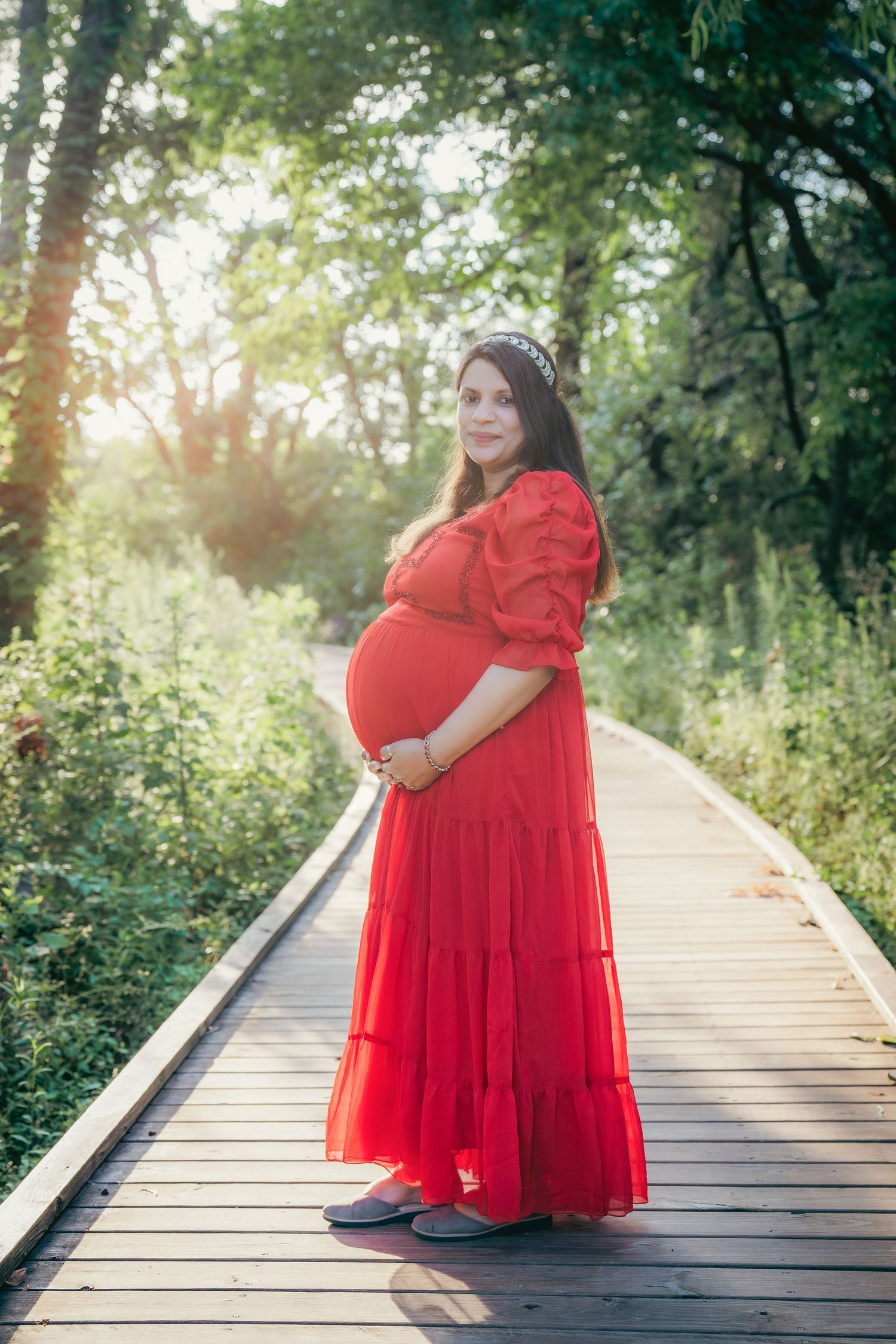 Maternity photoshoot on wooden boardwalk with flowing red dress in sunlit forest setting.