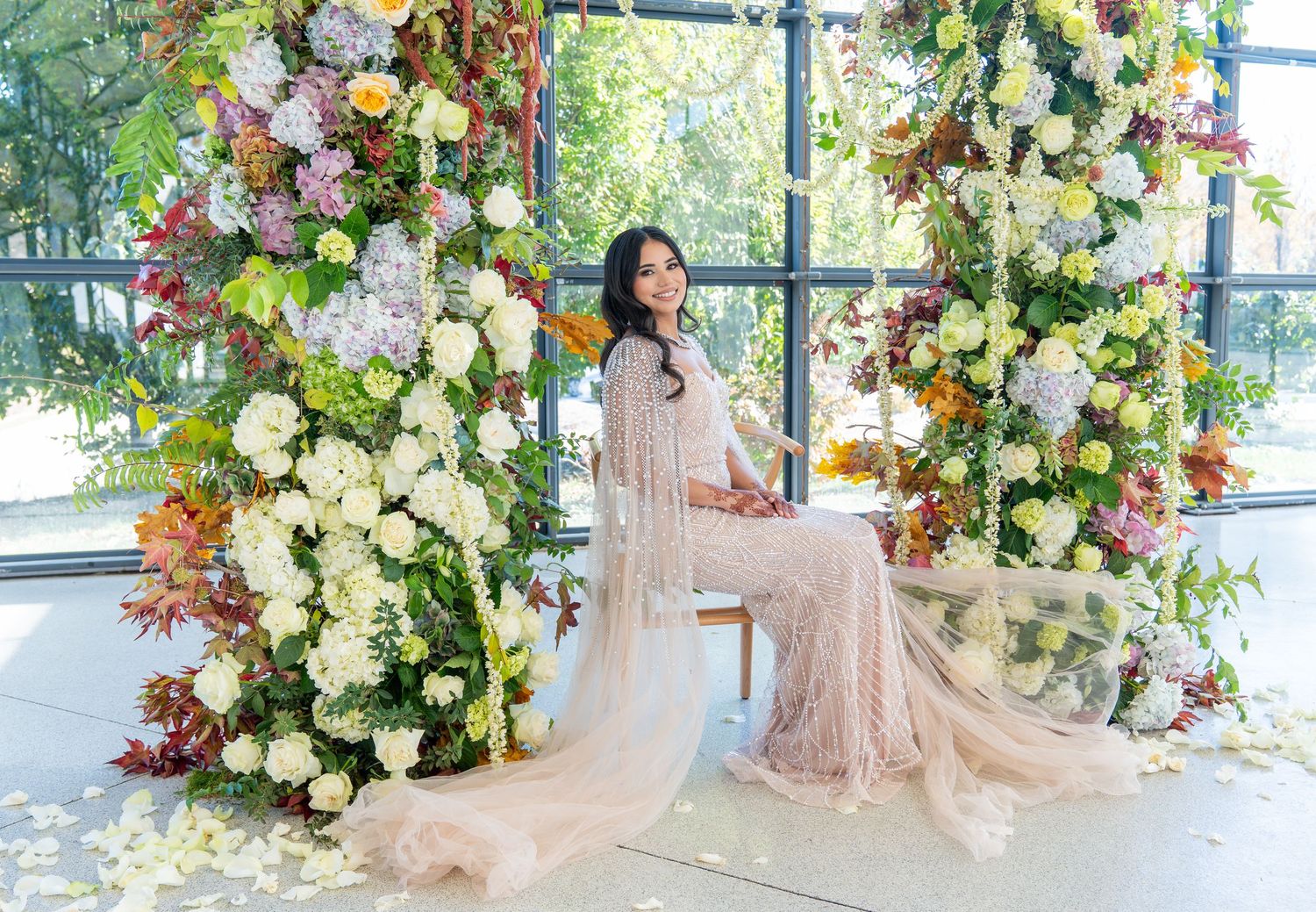 Bride in beige gown sits under elaborate floral arch with roses and mixed blooms.