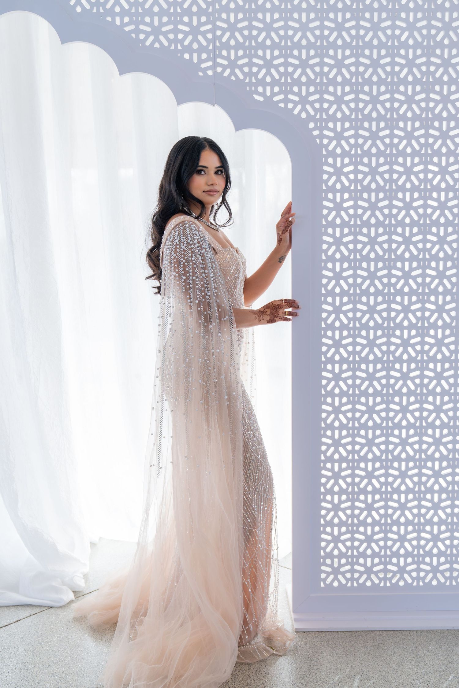 Bride in sheer beaded gown poses against geometric pattern wall.