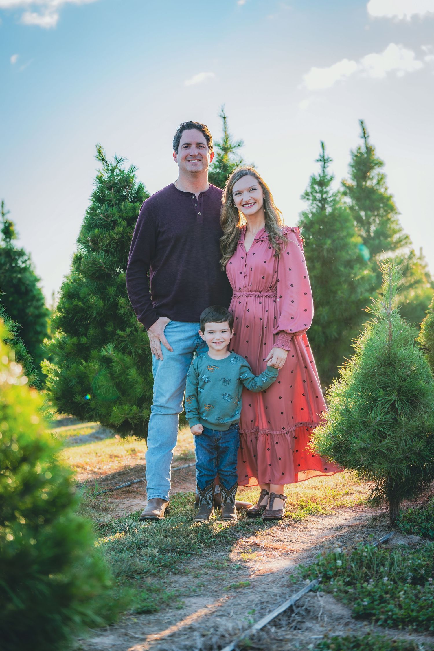 Family walking together among evergreen trees at a Christmas tree farm during golden hour.