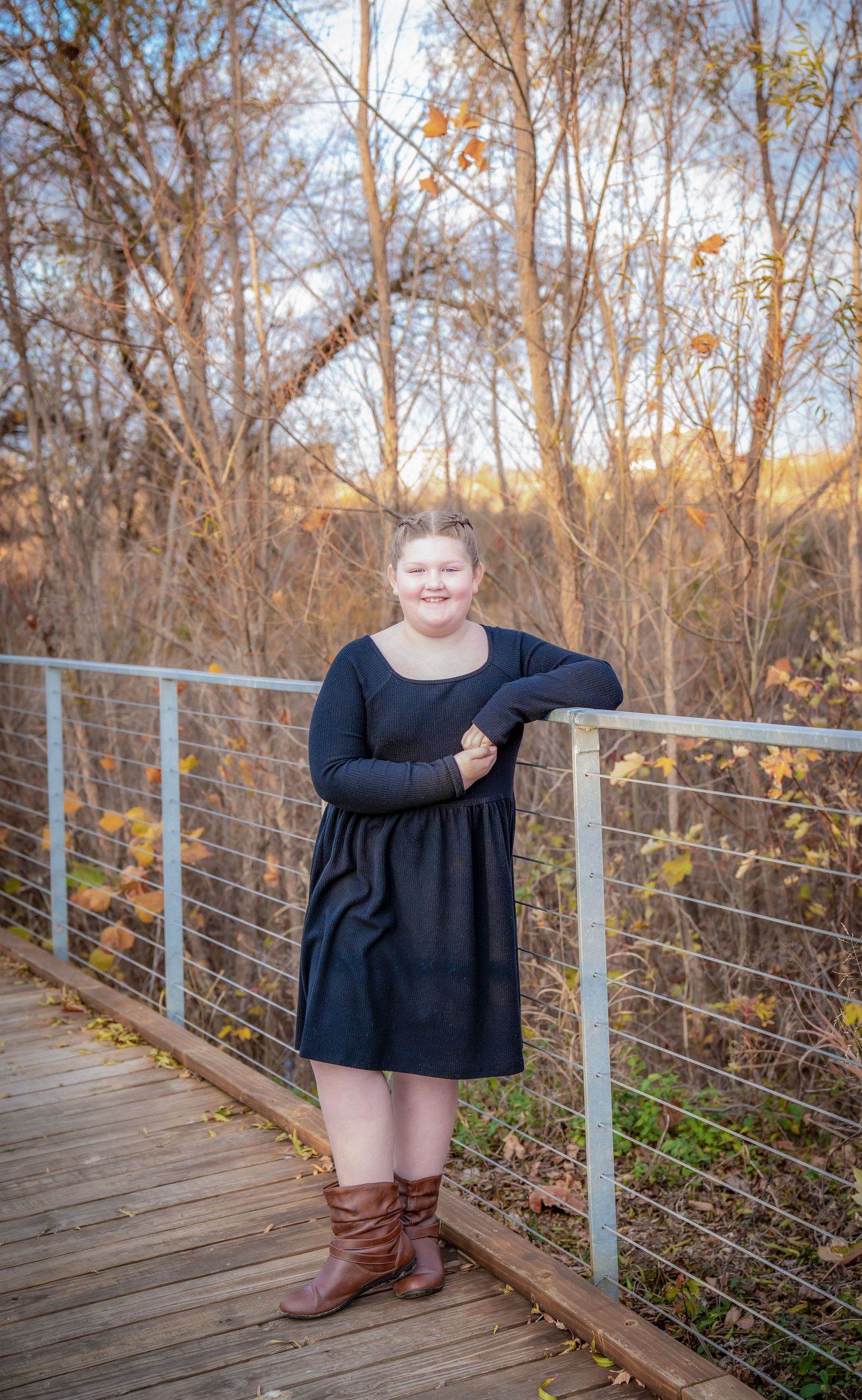 A person in a black dress poses on a wooden boardwalk surrounded by autumn trees.