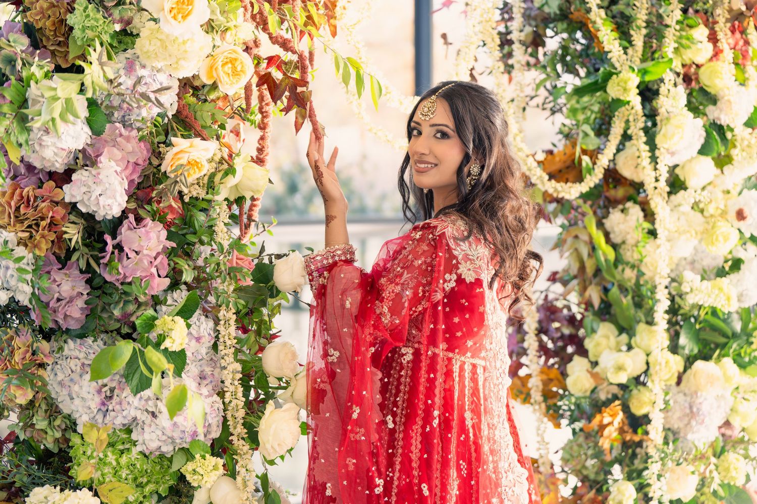 Bride in red lehenga surrounded by elegant floral decorations at Indian wedding venue.