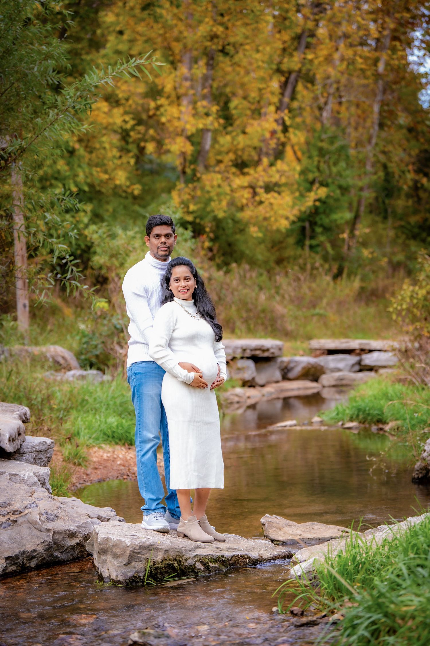 A couple sits together on rocky outcrops by a stream with fall colors in the background.