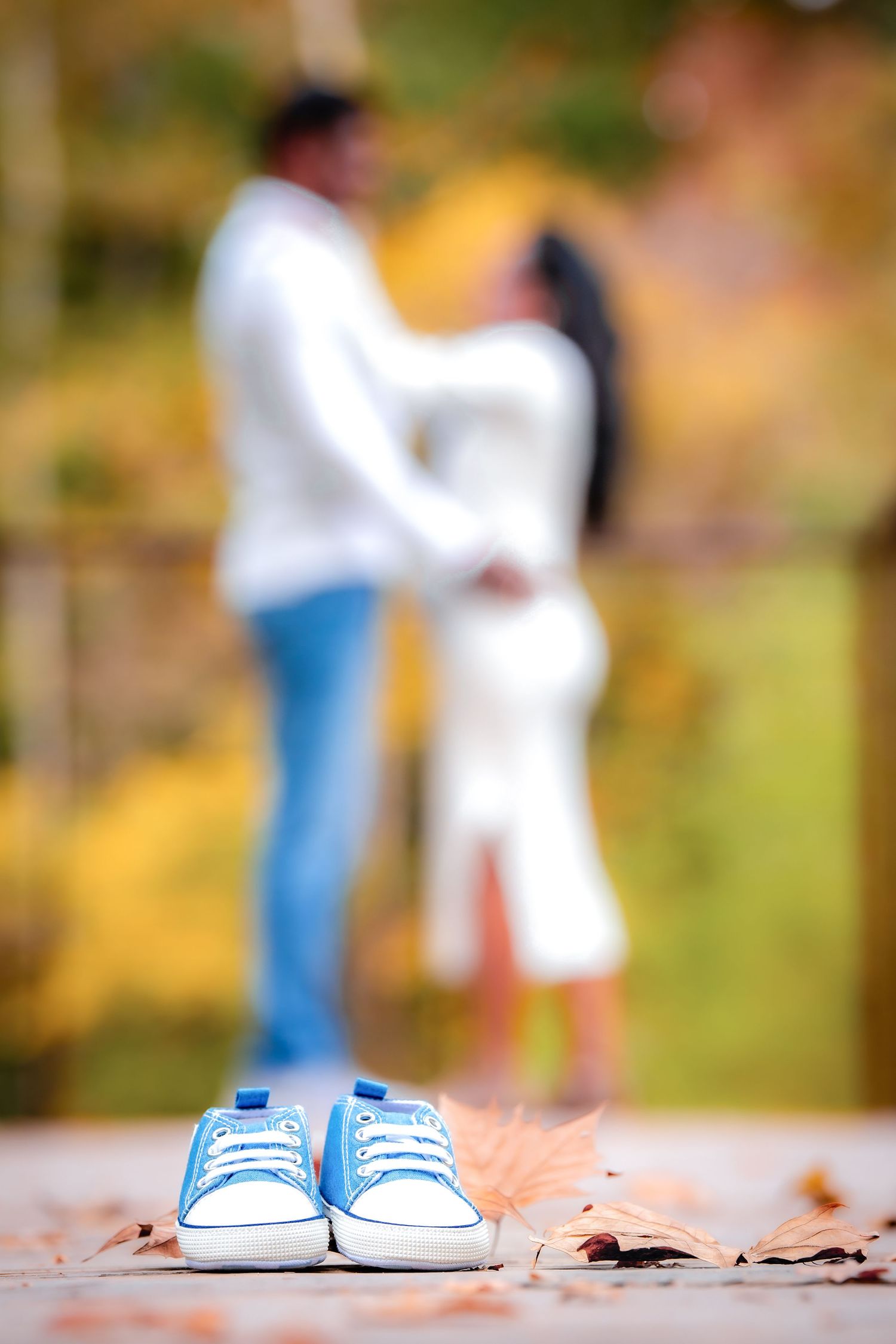 Small blue baby shoes in focus with blurred couple standing in autumn background.