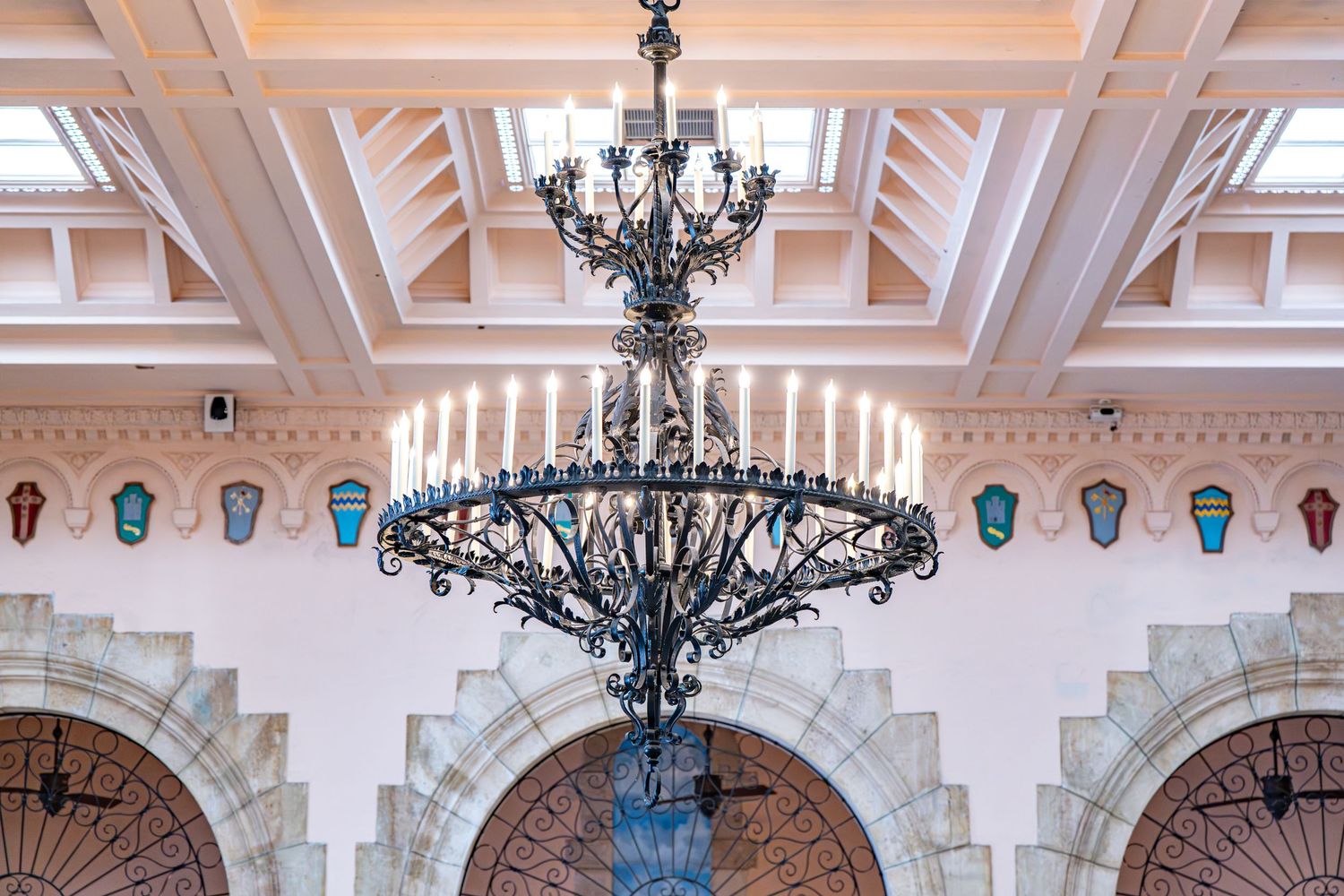 Ornate black iron chandelier hangs from a coffered ceiling with decorative turquoise masks mounted on the walls.