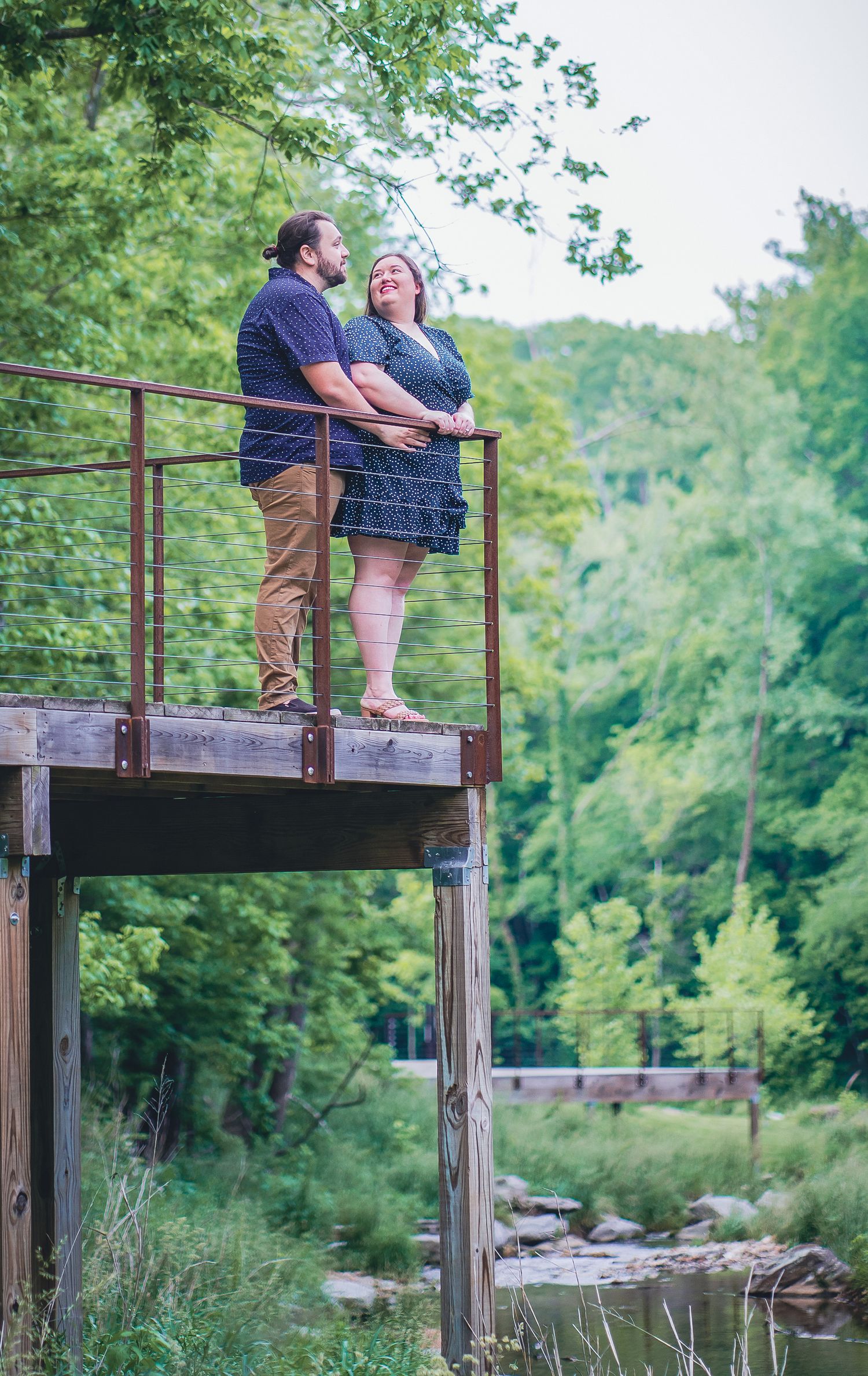 Couple sitting together on a wooden observation deck overlooking a scenic creek surrounded by lush green trees.
