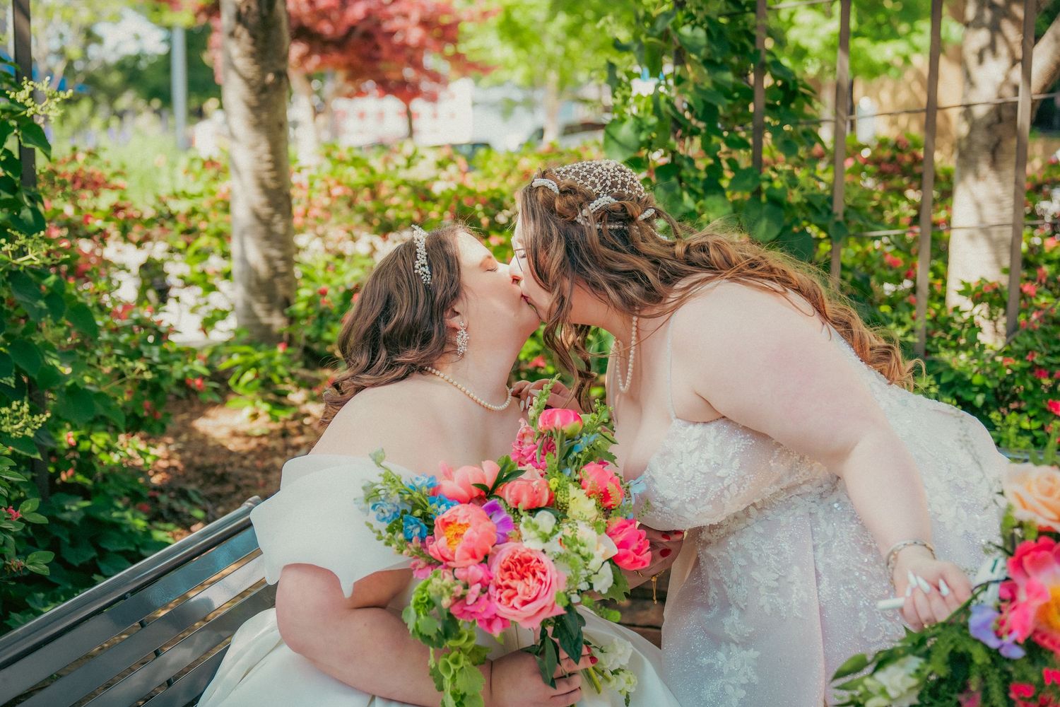 Two people in white wedding dresses hold bouquets in a lush garden setting.