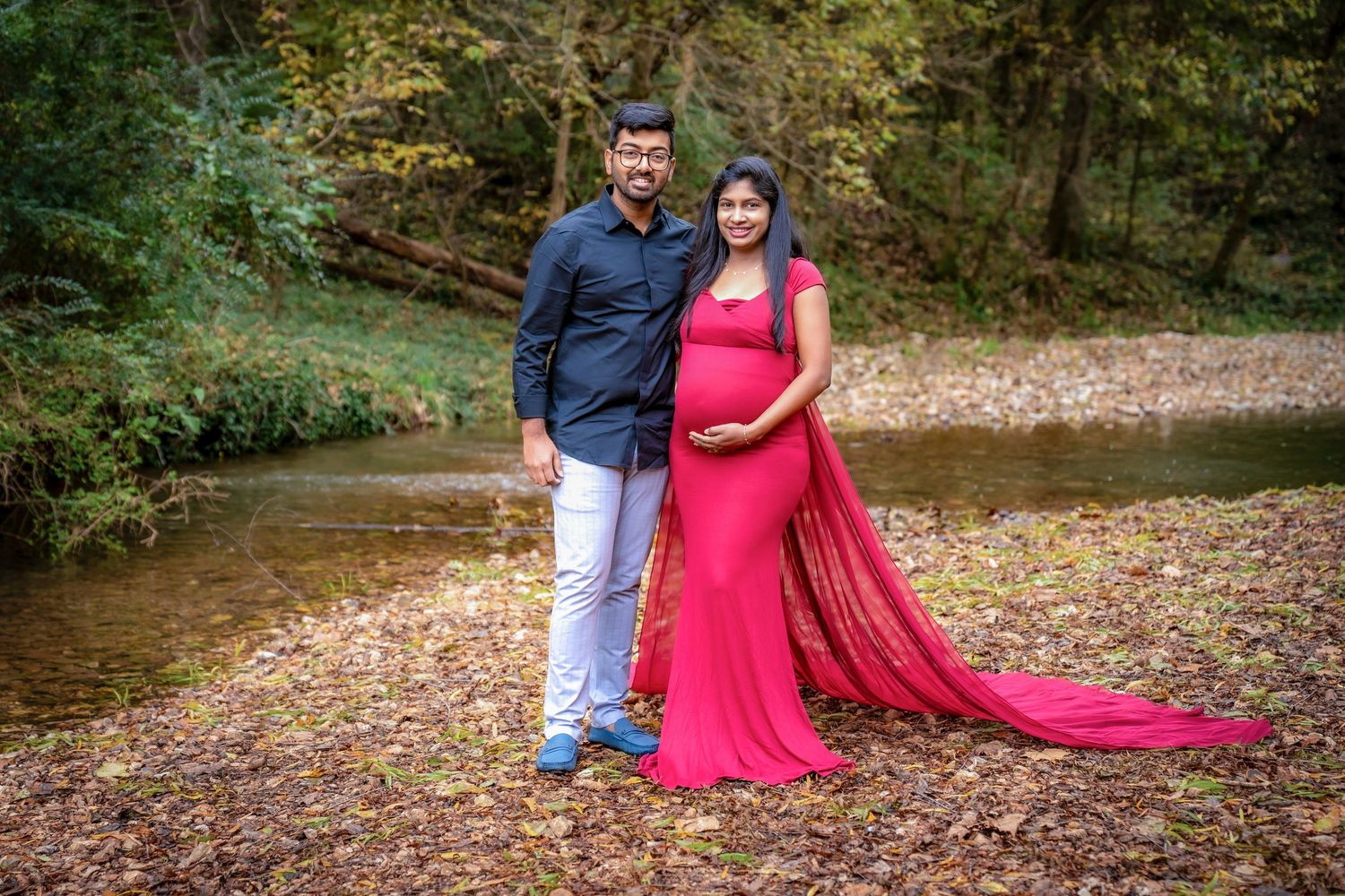 A couple poses together in a wooded setting with fall foliage and natural lighting.