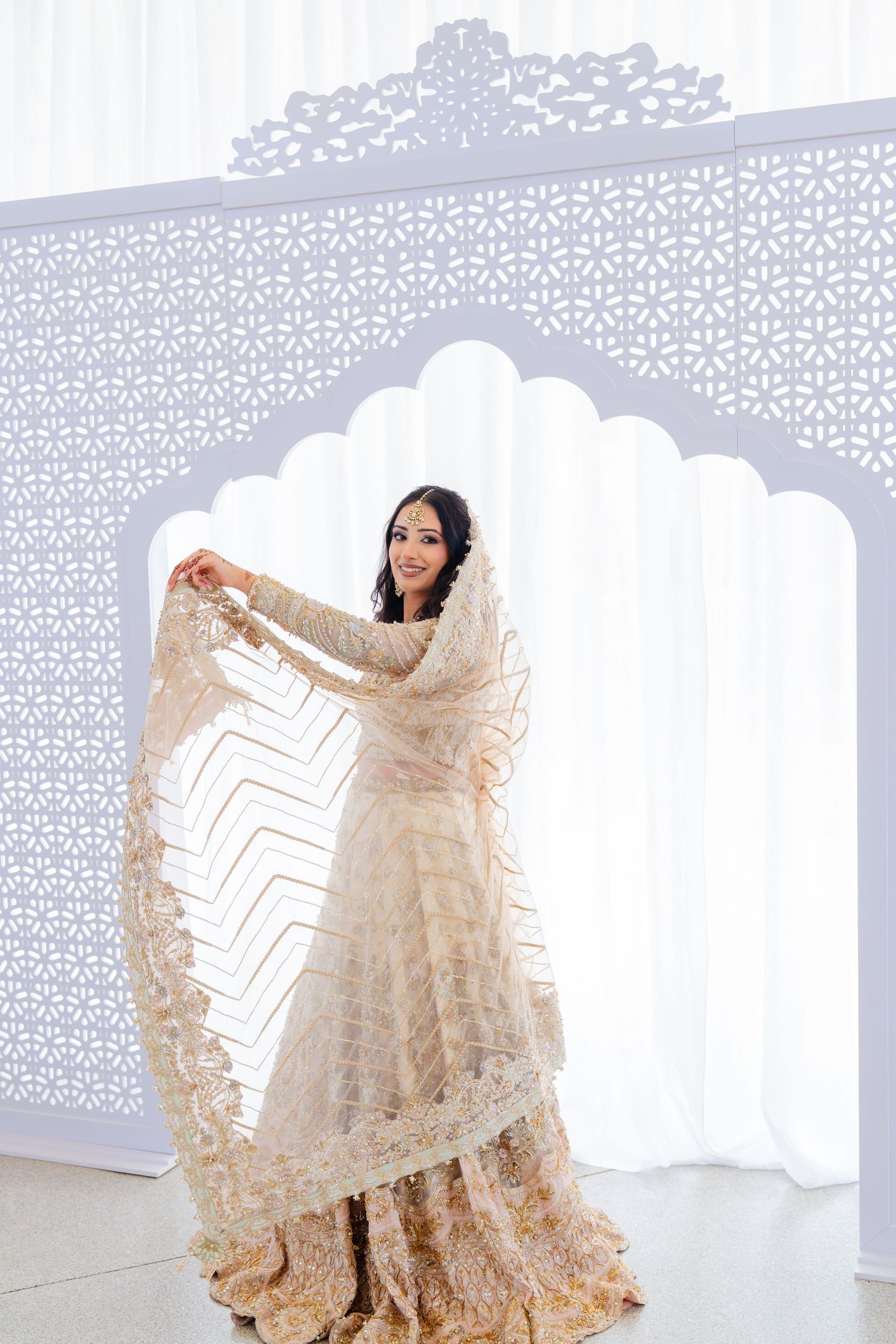 Bride in cream lehenga holding dupatta dramatically against geometric backdrop.