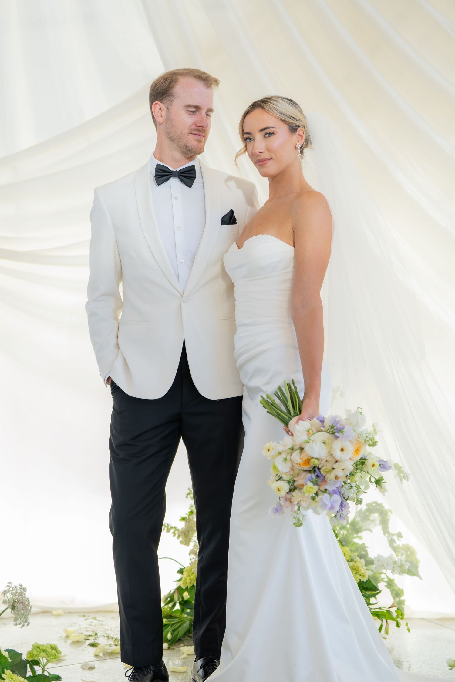 Bride and groom stand together in formal attire under white tent with floral decor.