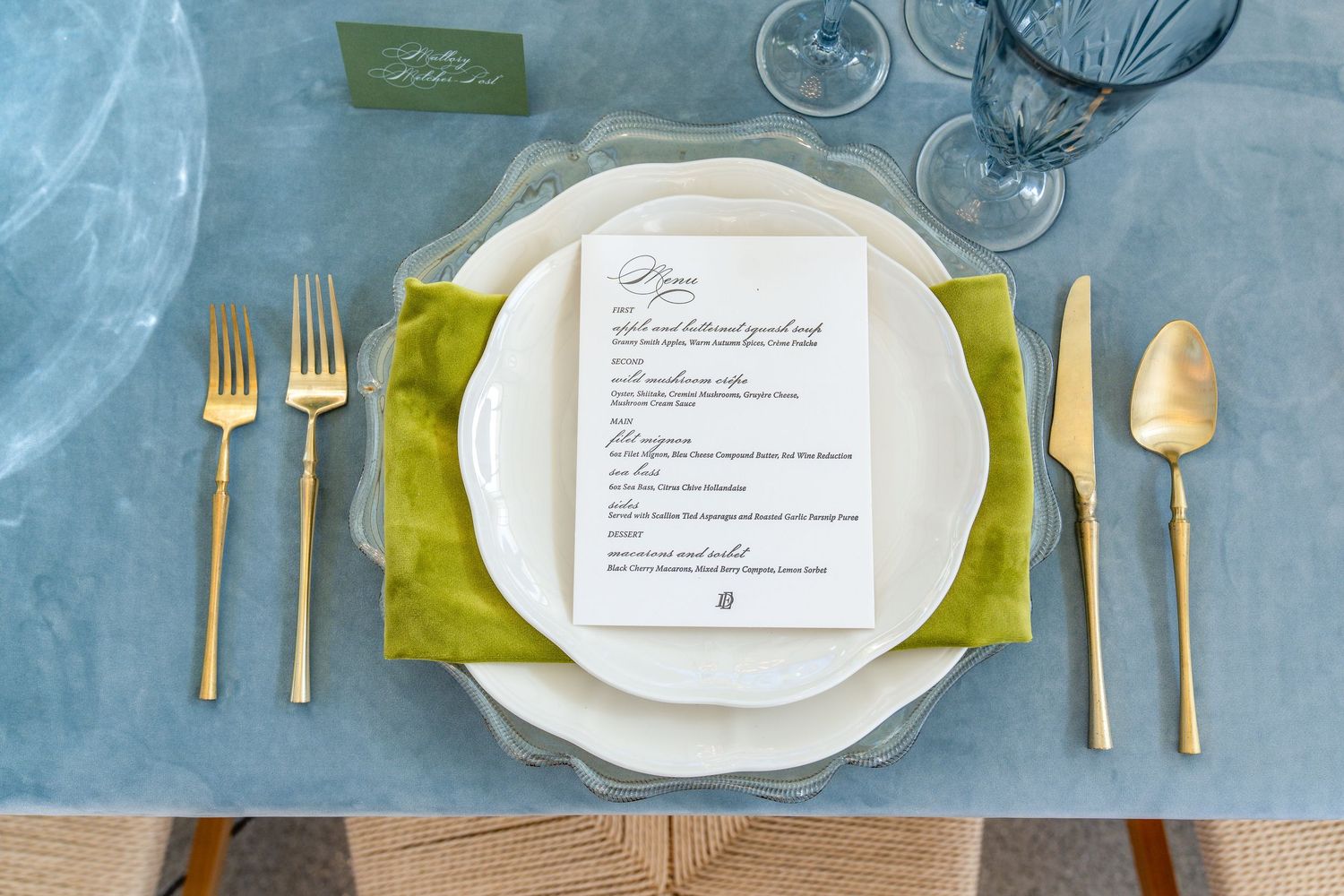 Elegant place setting with gold flatware, white plate, and green napkin on blue tablecloth.