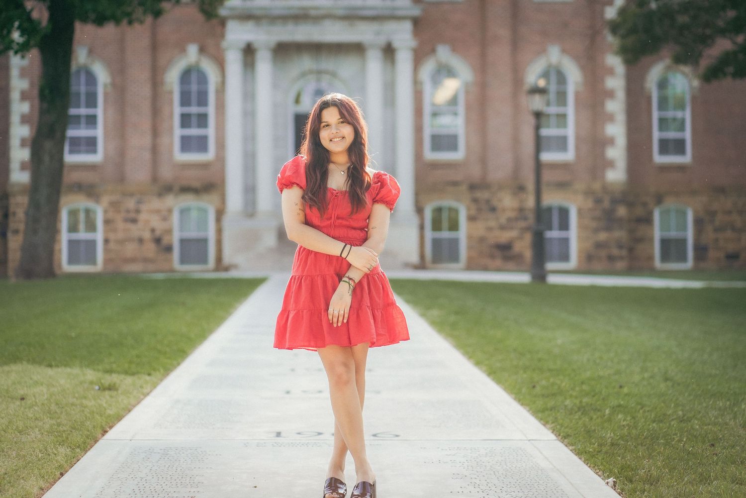 Graduate in red dress and cap walking along pathway with brick buildings in background.