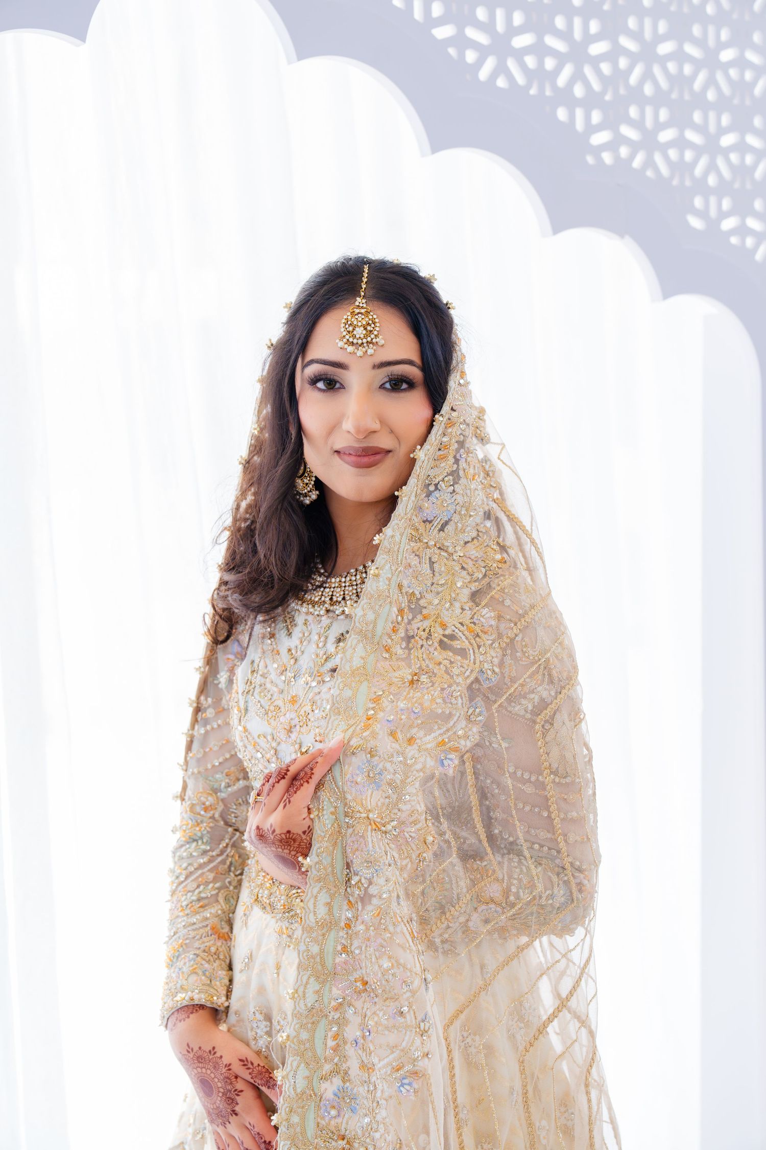 South Asian bride wearing intricate beige and gold lengha with jewelry against white backdrop.