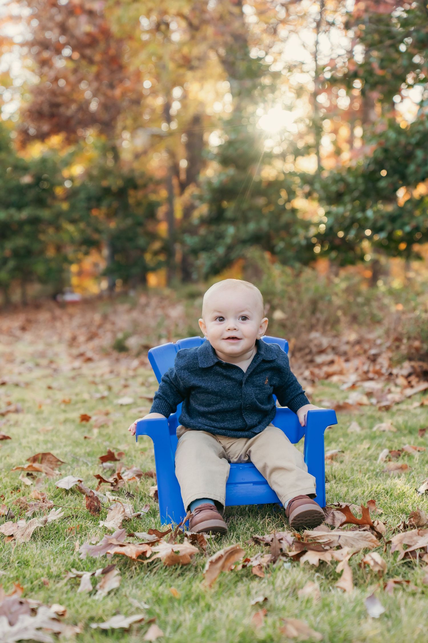 Baby sitting in small blue chair surrounded by fallen autumn leaves on grass.