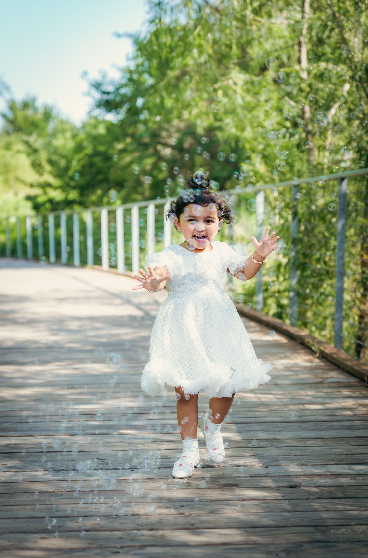 A toddler in a white lace dress and shoes plays joyfully on a wooden boardwalk surrounded by greenery.