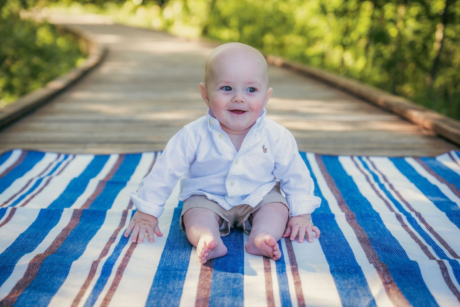 Baby in white outfit playing on blue striped blanket outdoors.