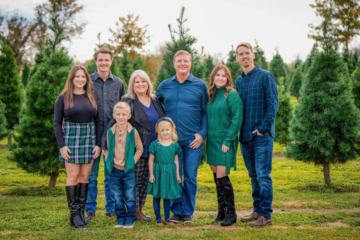 A multi-generational family portrait among evergreen trees wearing coordinated casual attire.