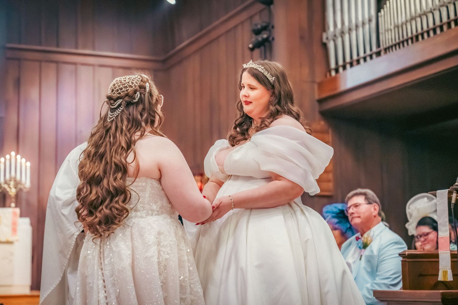 Two people in flowing white wedding dresses embrace during a church ceremony.