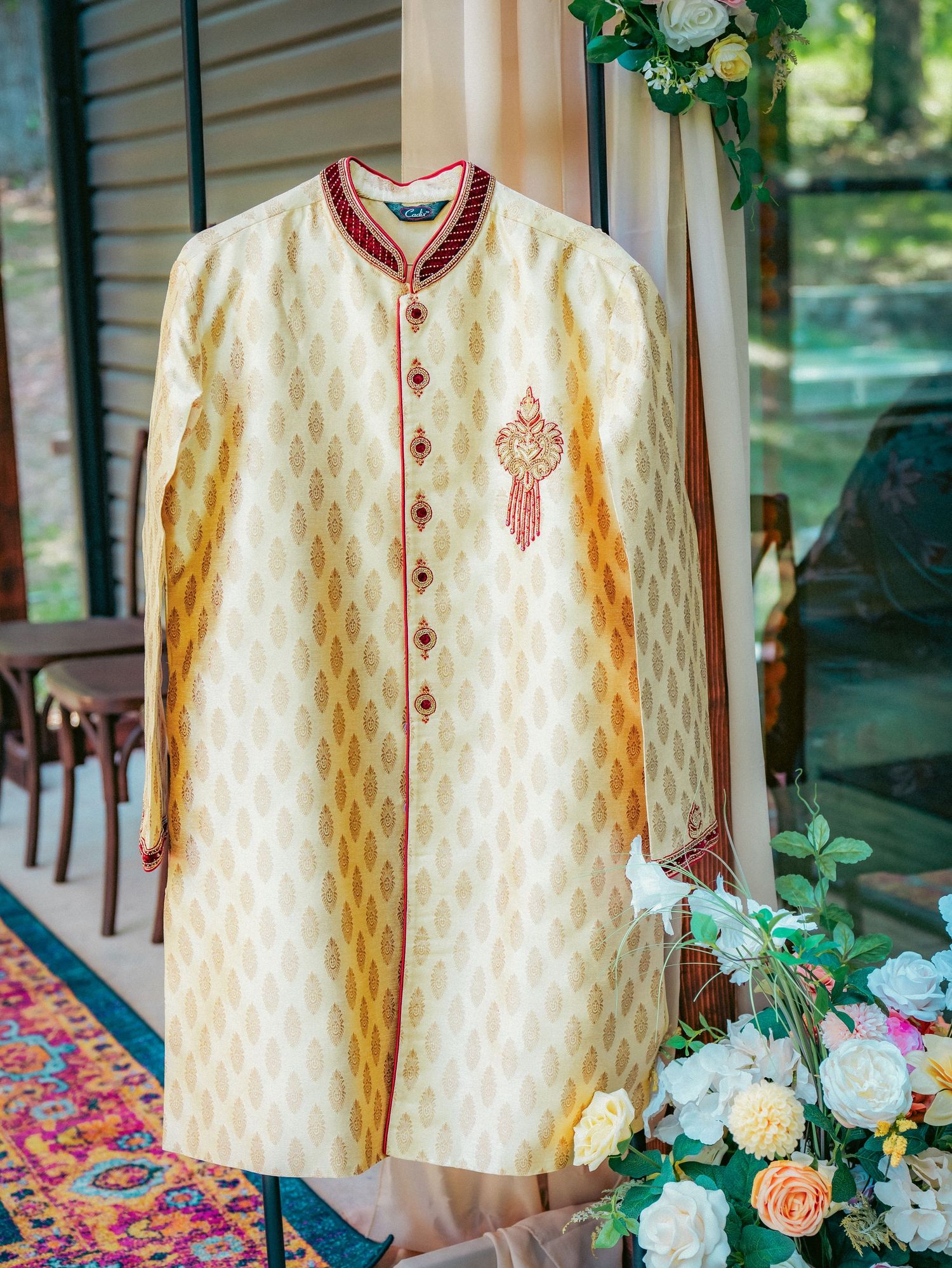 An ivory and gold sherwani with red accents and detailed embroidery displayed among floral decorations.