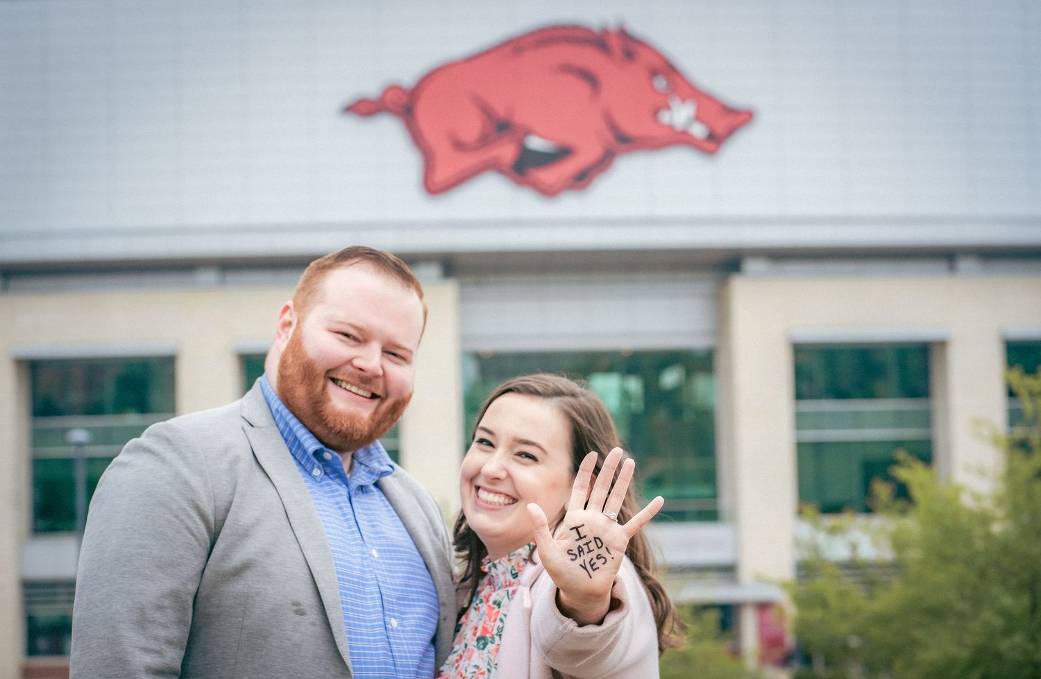 Happy couple showing engagement ring with Razorback mascot visible in background.