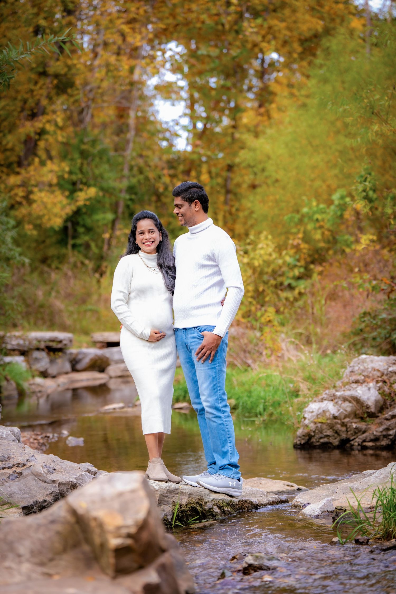 Two people pose playfully on rocks near a stream with autumn trees creating a golden backdrop.