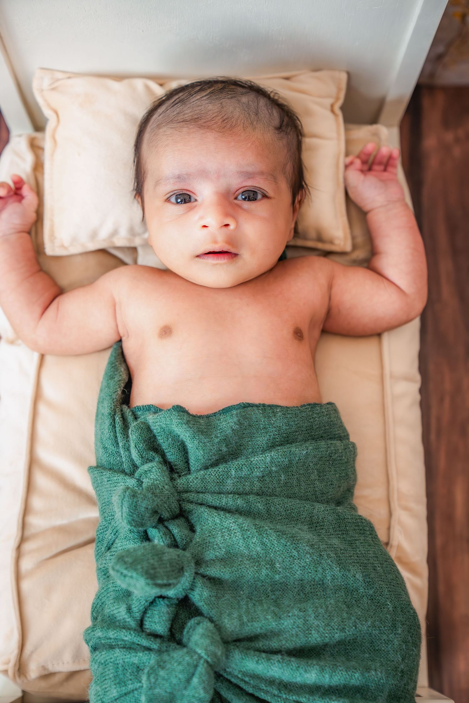 Newborn baby wrapped in green blanket lies on cream colored cushions.