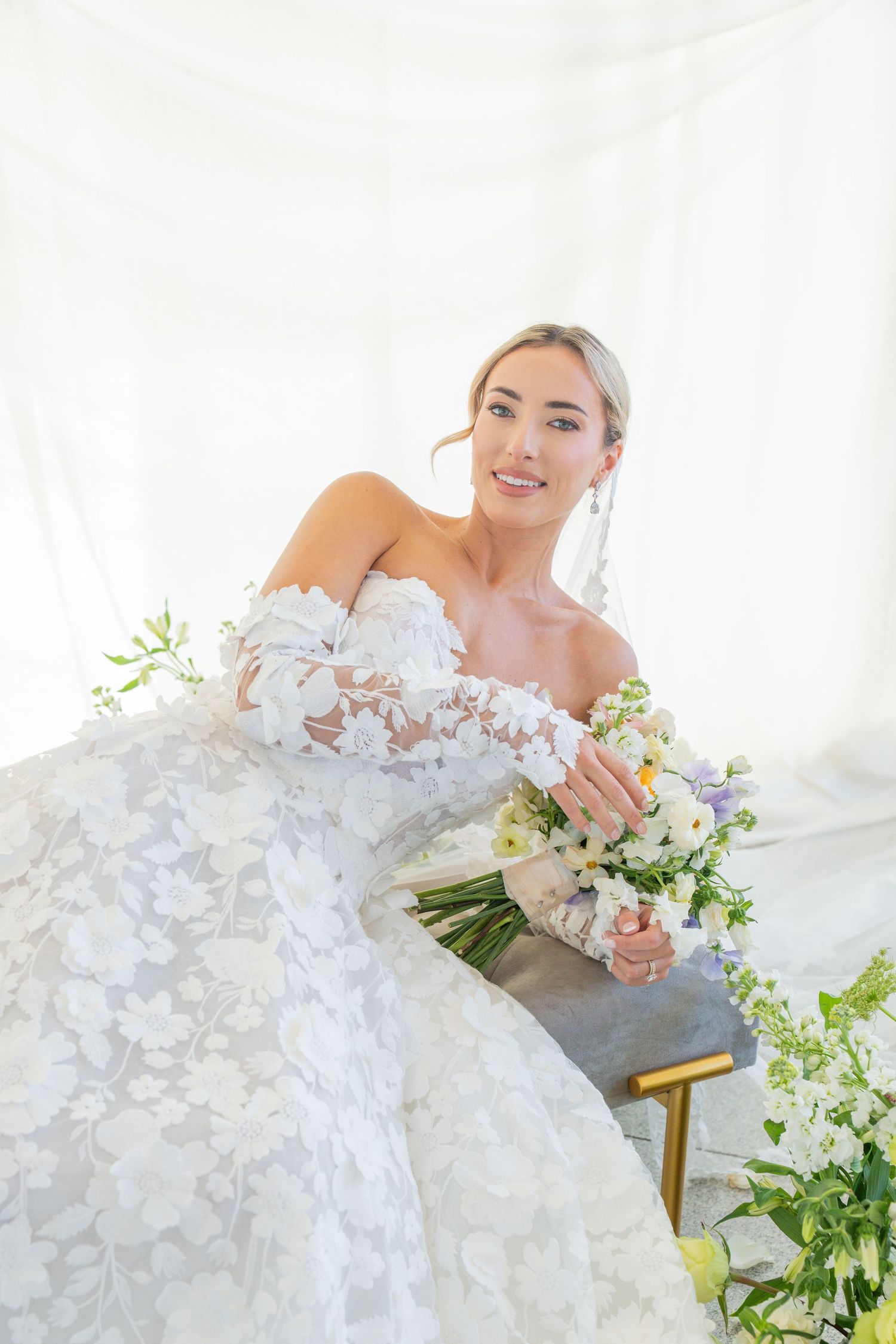 Bride in strapless lace wedding dress poses with spring bouquet against white backdrop.