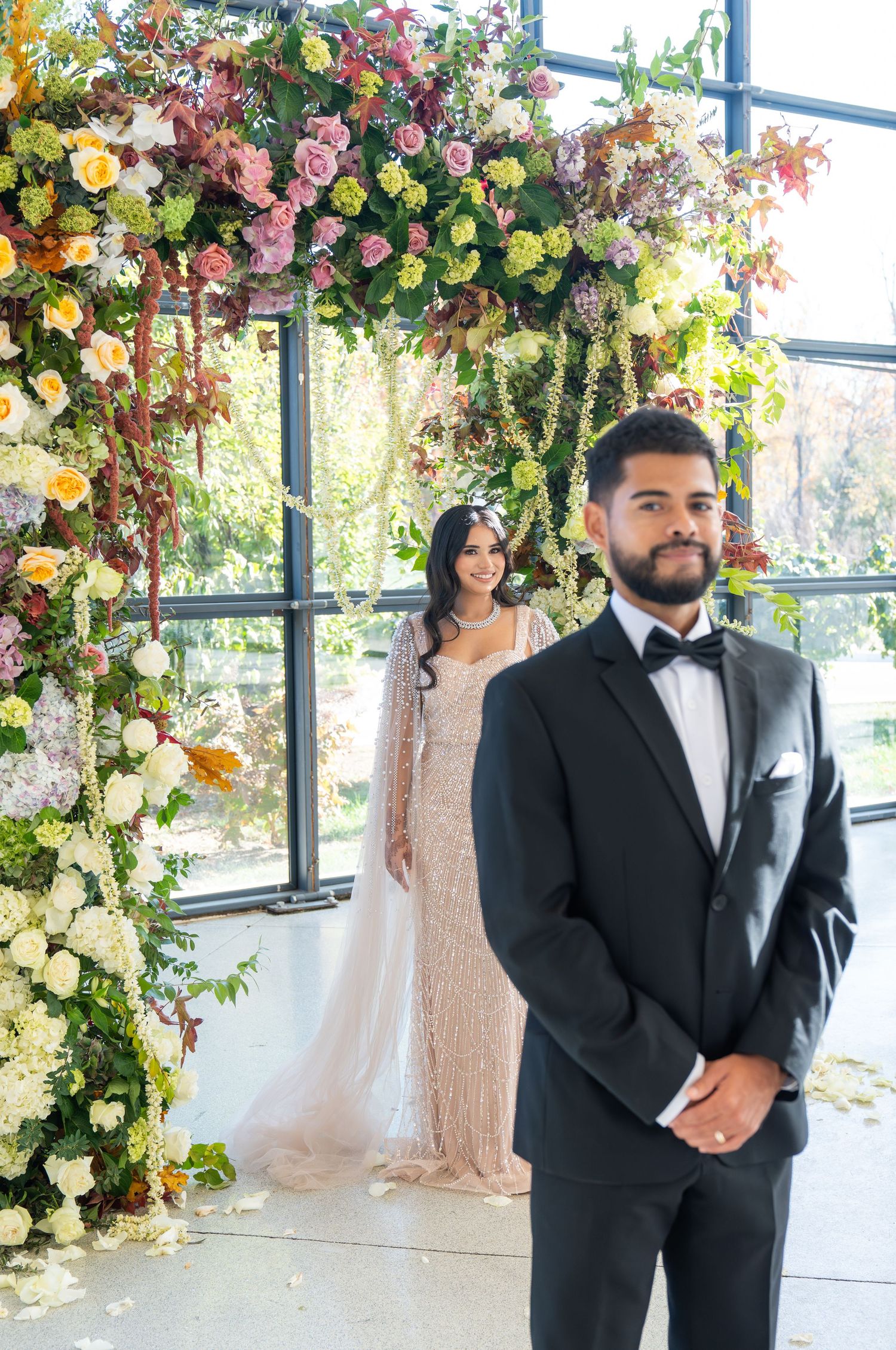 A first look wedding moment captured in a sunlit greenhouse decorated with colorful floral arrangements.