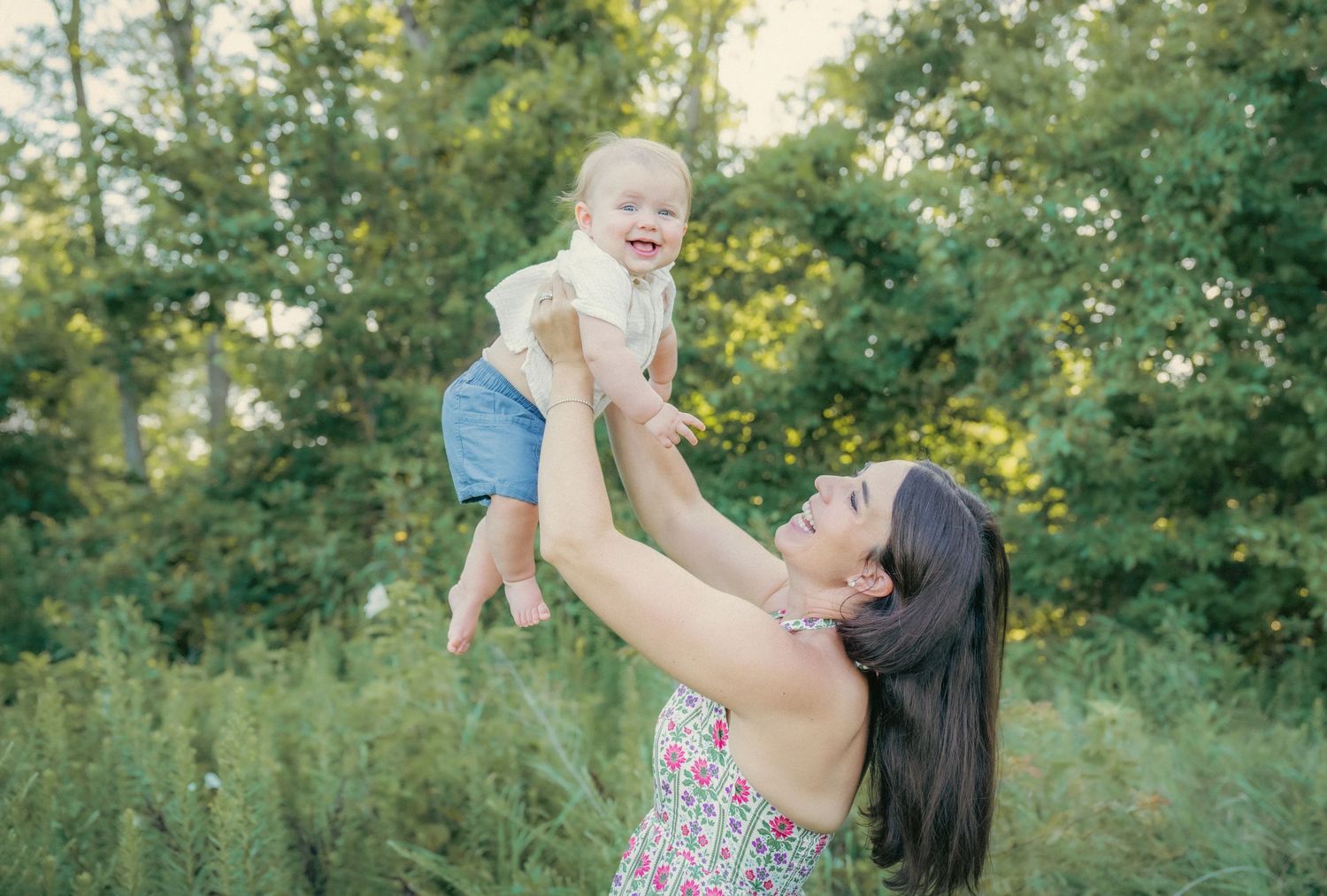 Mother and baby sharing joyful moments in a sunny outdoor setting with natural greenery.