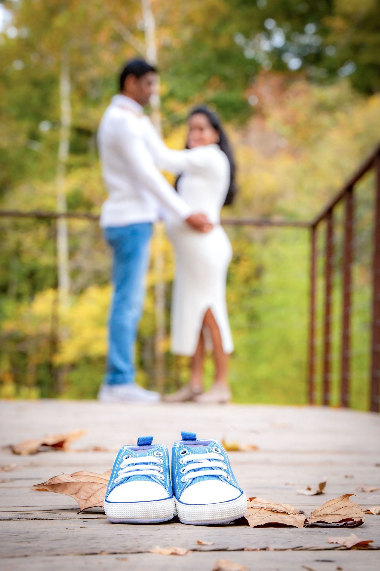 A pair of blue baby shoes sits on a wooden deck with autumn leaves while a couple embraces in the background.
