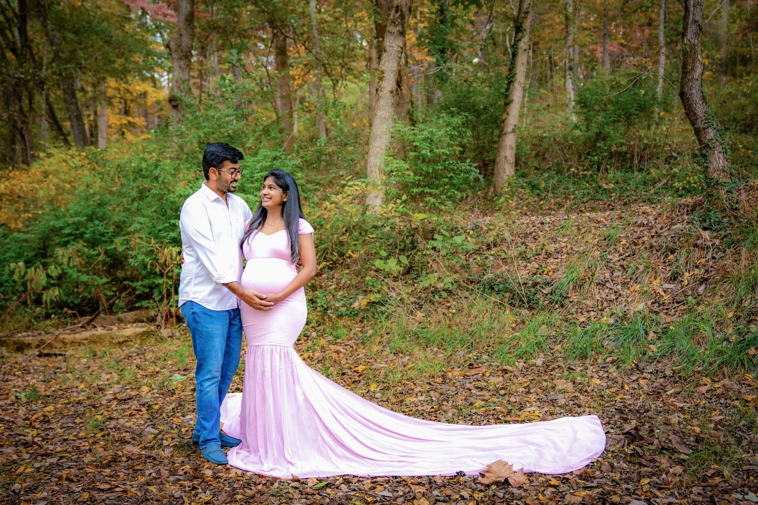 A flowing pink maternity dress creates an elegant silhouette against autumn trees and fallen leaves.