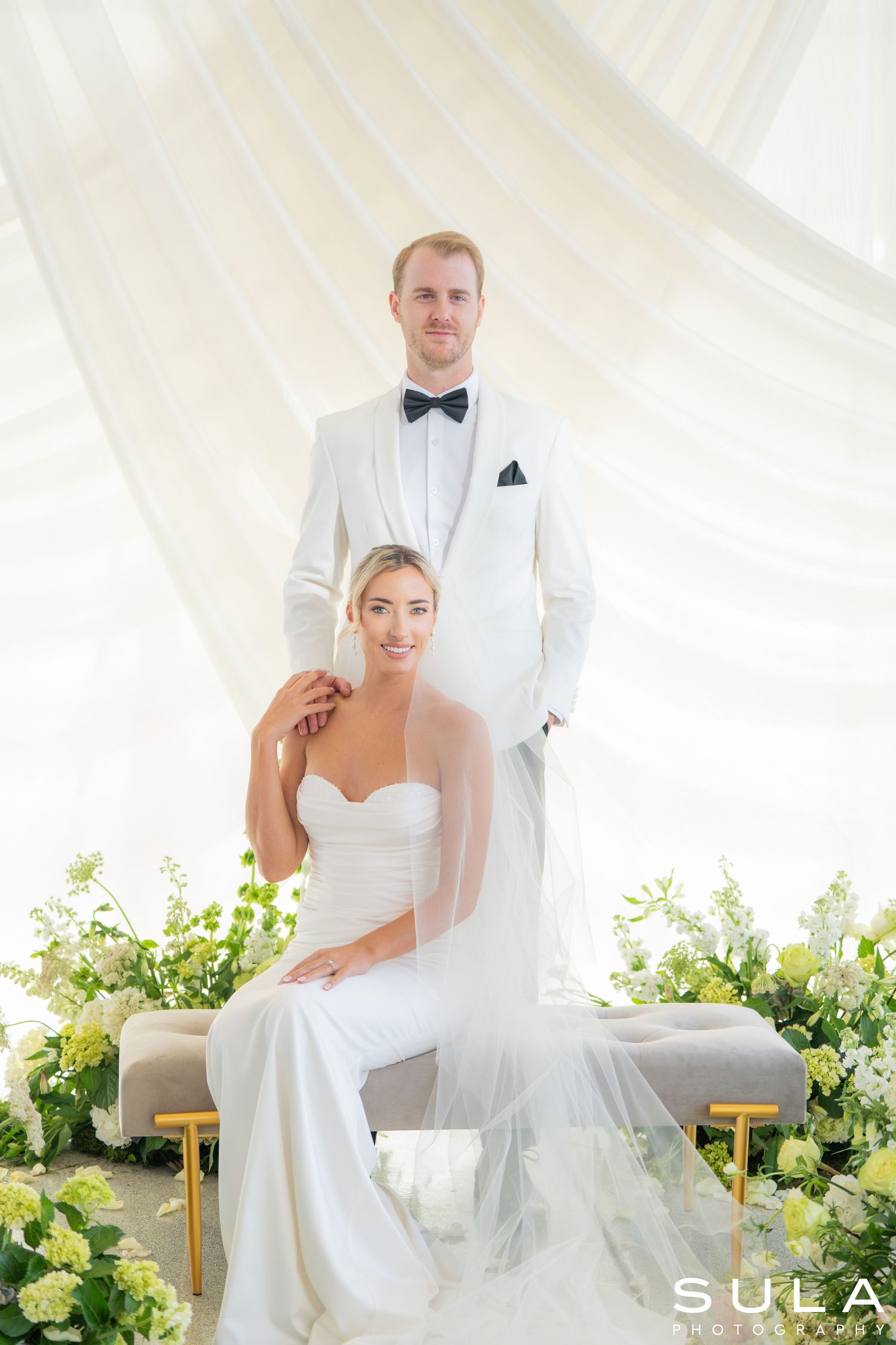 Elegant wedding portrait with white floral arrangements and flowing fabric backdrop.