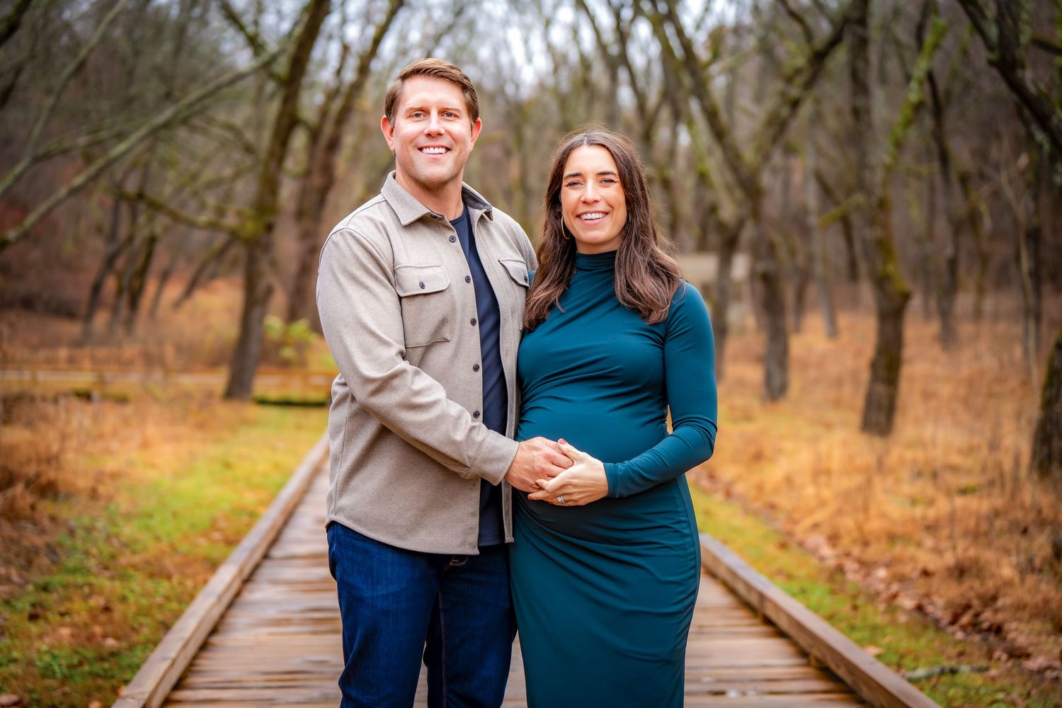 A couple stands together on a wooden boardwalk surrounded by bare autumn trees.